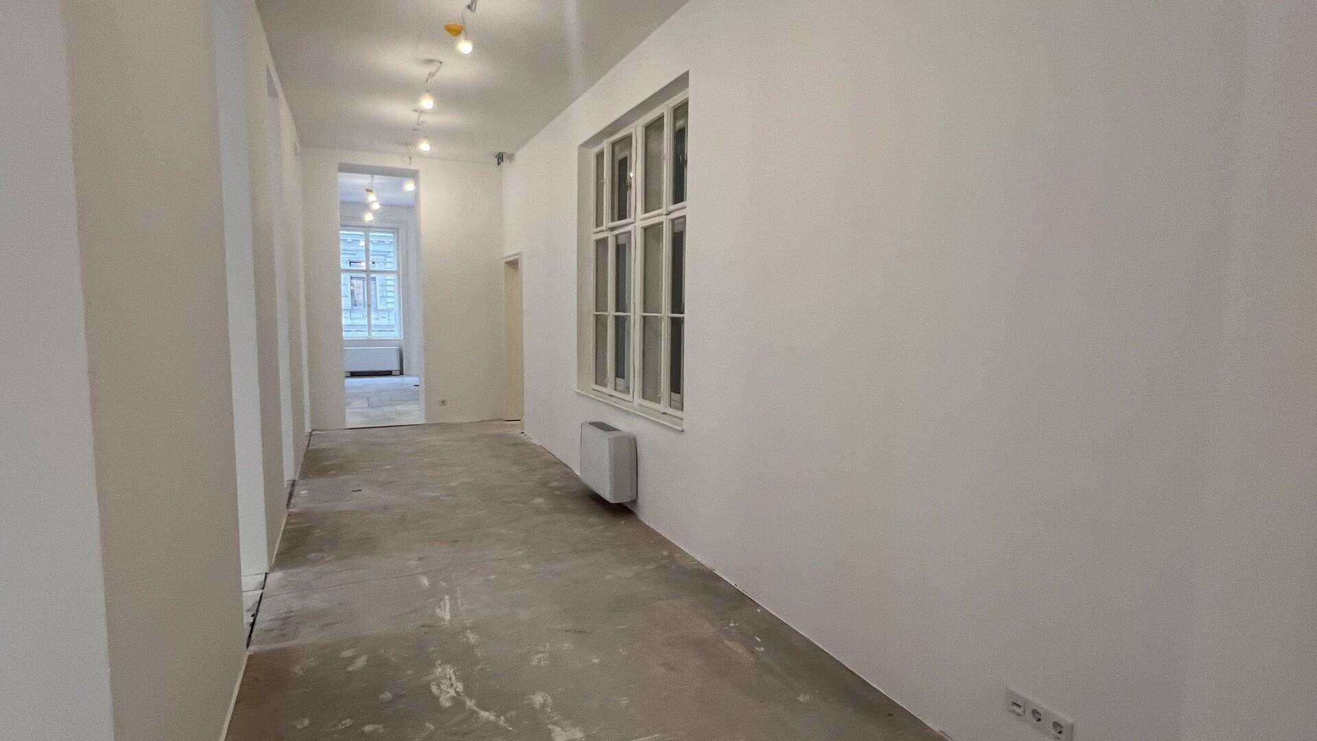 Langer Flur mit weiß gestrichenen Wänden und einem Fenster, das Tageslicht hereinlässt.