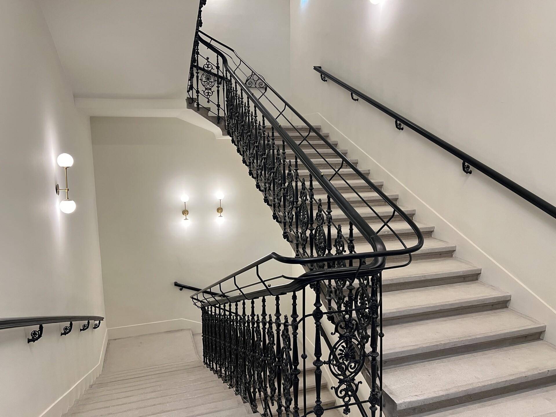 Elegantes Treppenhaus mit schmiedeeisernem Geländer und hellen Stufen, beleuchtet von Wandlampen.