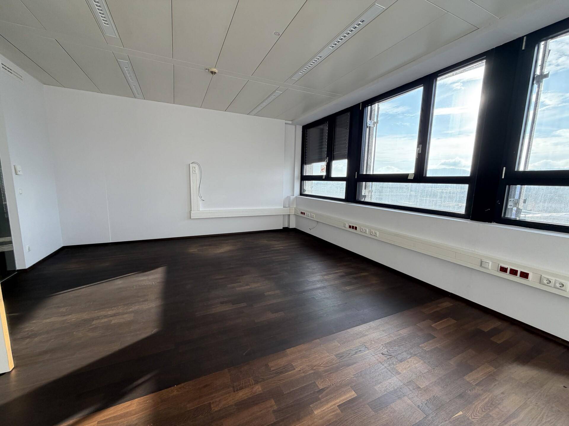 Leeres Büro mit großen Fenstern, dunklem Holzboden und viel natürlichem Licht.