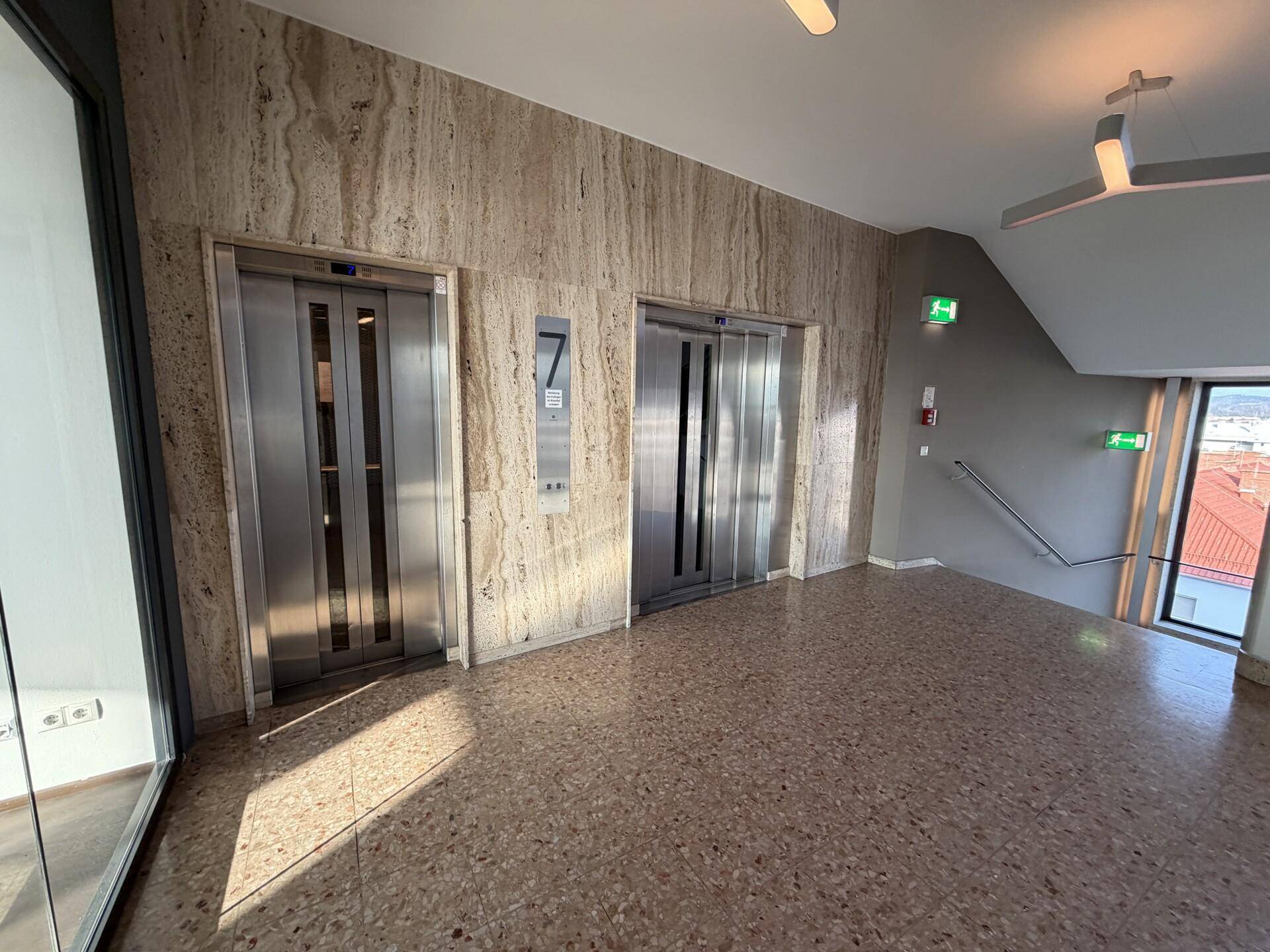 Heller Flur mit zwei Aufzügen, Natursteinwänden und Terrazzoboden in einem modernen Gebäude.