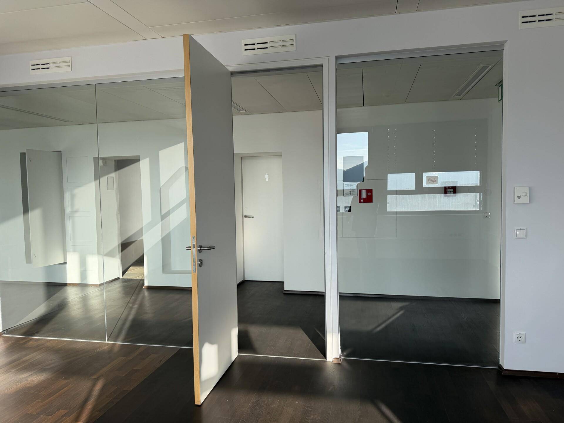 Moderner Büroraum mit Glaswänden, Holzboden und einer offenen Tür, die den Zugang ermöglicht.