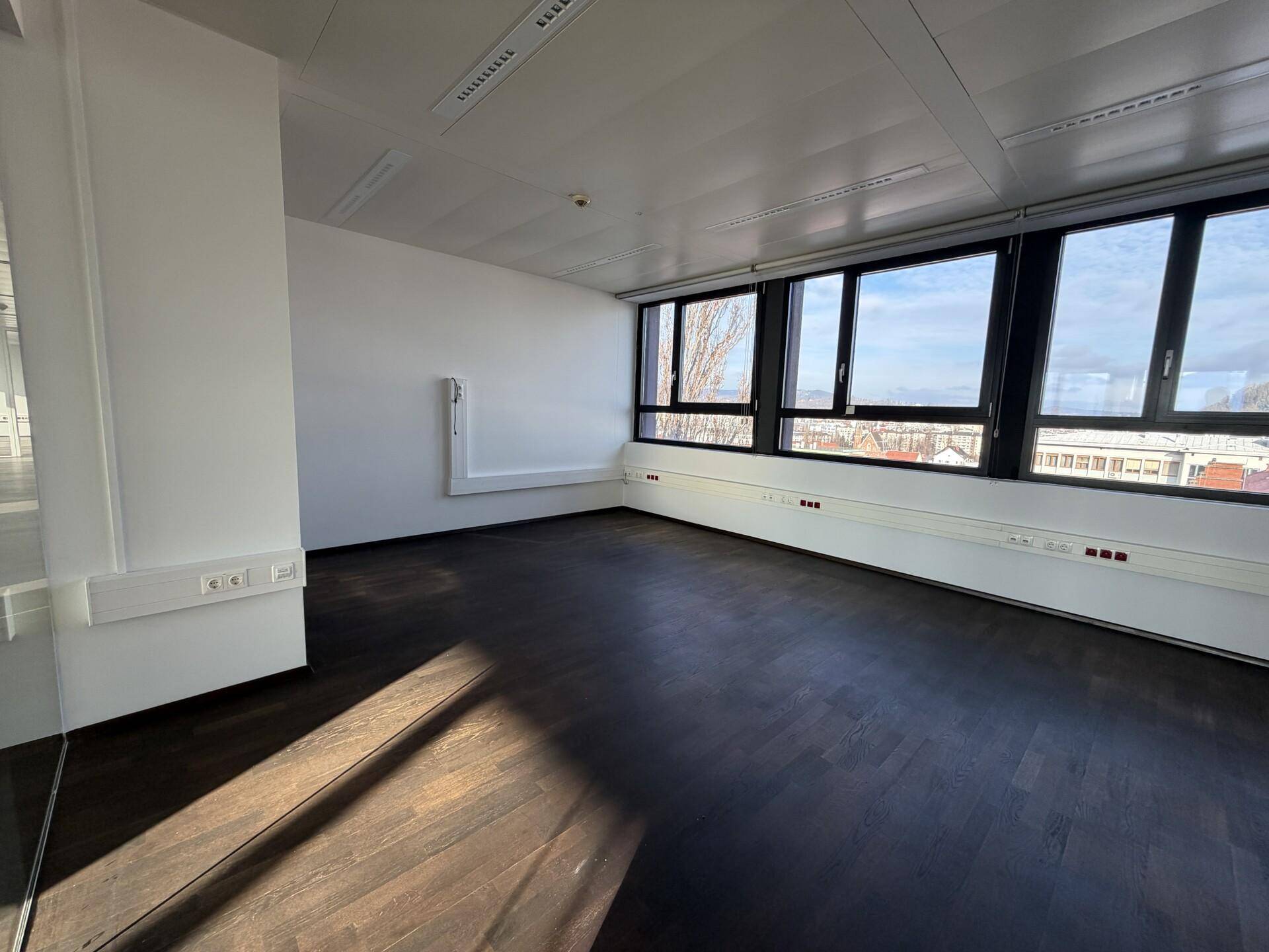 Geräumiges, leeres Büro mit dunklem Holzboden und einer Fensterfront, die viel Licht hereinlässt.