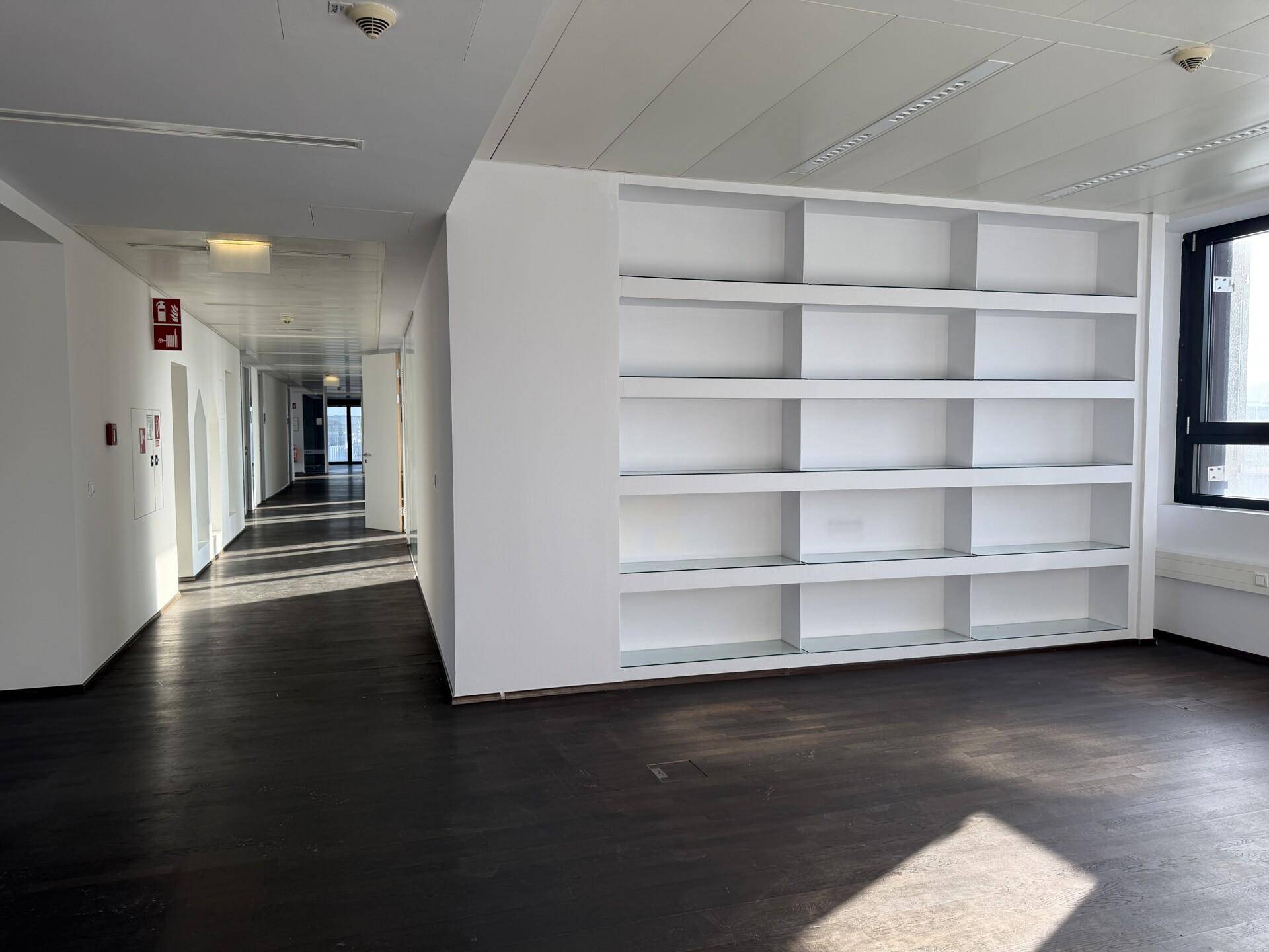 Geräumiger Büroflur mit dunklem Holzboden, weißen Wänden und einem großen, leeren Regal.