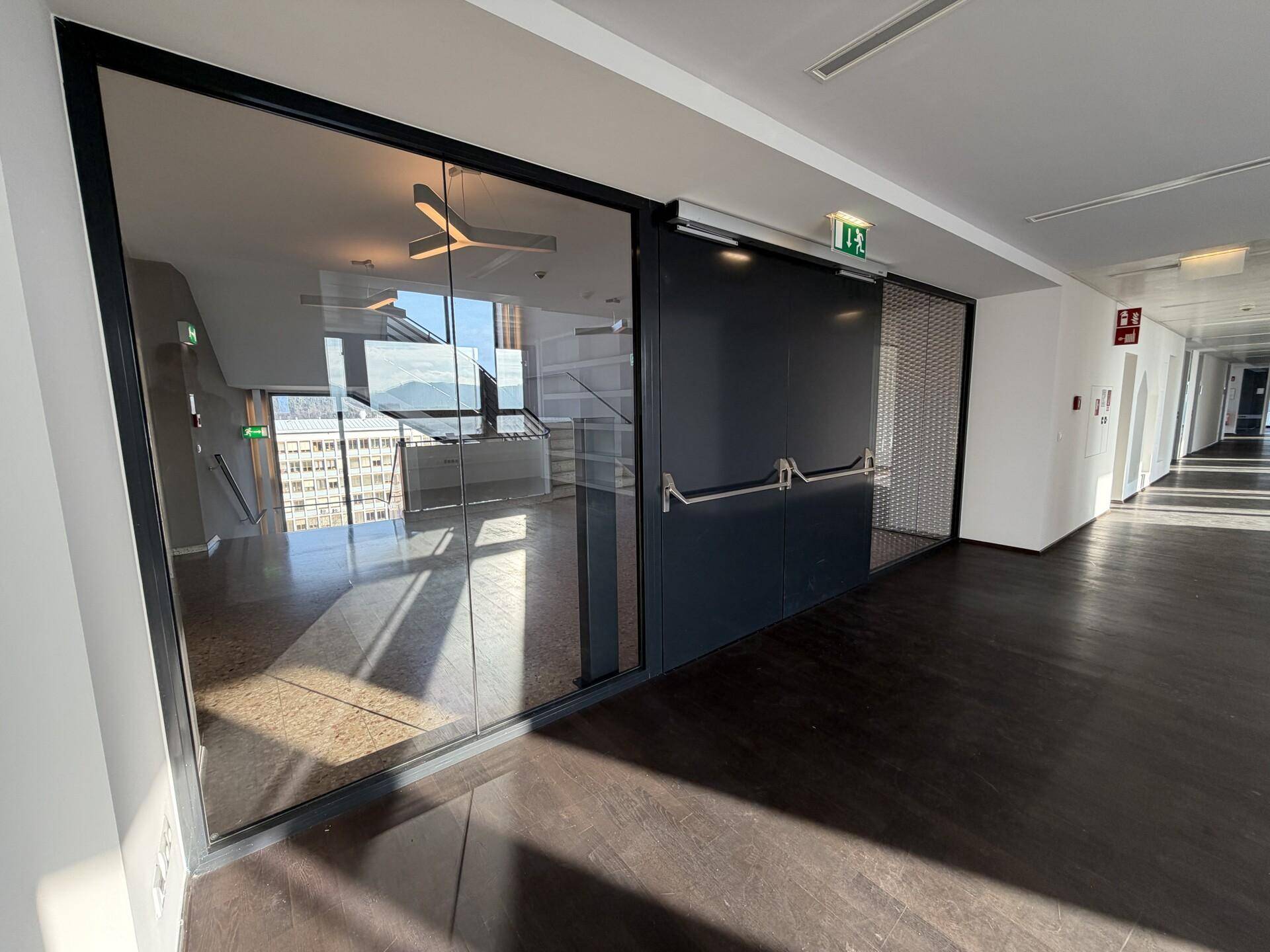 Moderner Büroflur mit Glaswänden, dunklem Holzboden und Blick in einen hellen Büroraum.