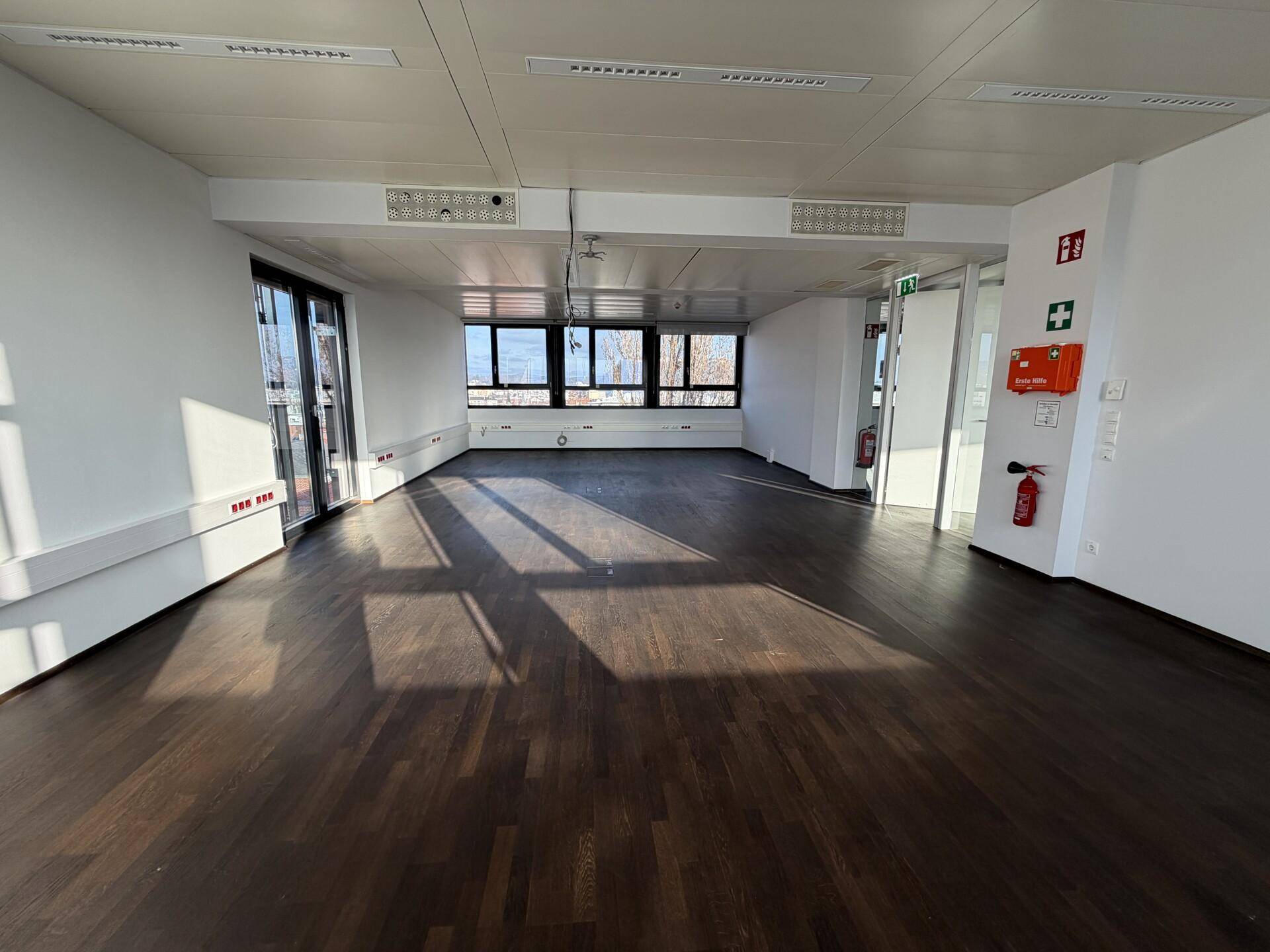 Großes, leeres Büro mit dunklem Holzboden, weißen Wänden und viel Tageslicht.