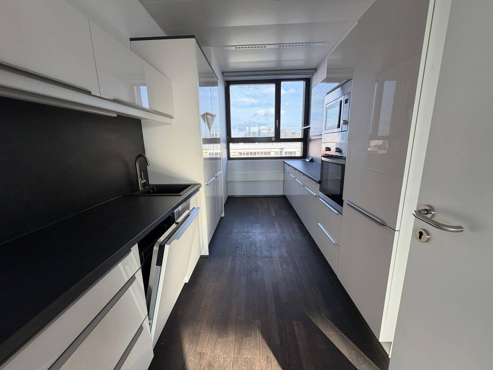 Moderne, schmale Küche mit weißen Hochglanzschränken, dunkler Arbeitsplatte und Fensterblick.
