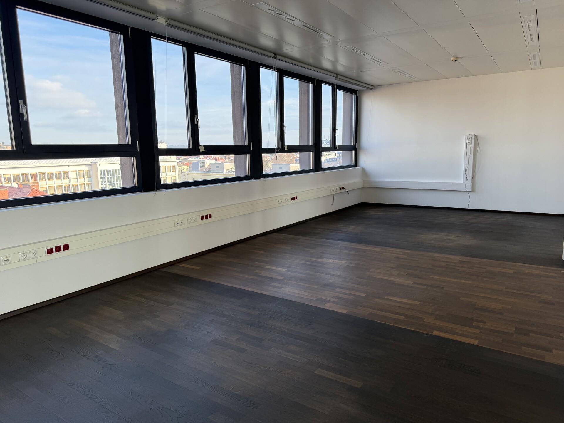 Leeres Büro mit dunklem Holzboden und einer langen Fensterfront, die den Raum erhellt.