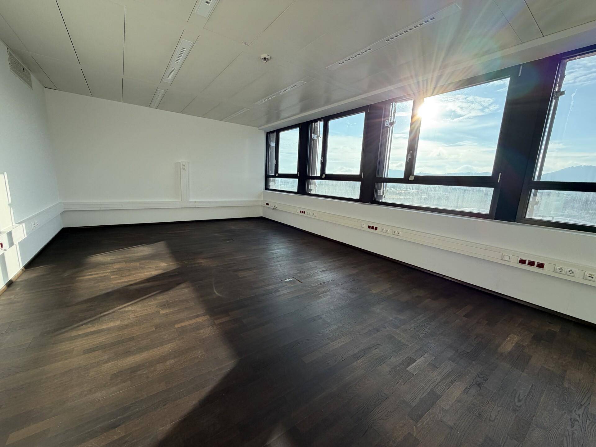 Geräumiges, leeres Büro mit dunklem Holzboden und einer breiten Fensterfront, die viel Licht spendet.