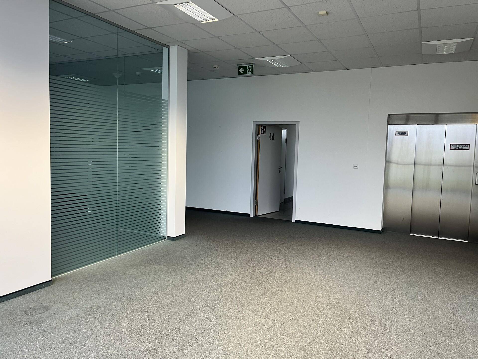 Moderner Bürobereich mit Teppichboden, Glaswand und Aufzug, bereit zur Einrichtung.