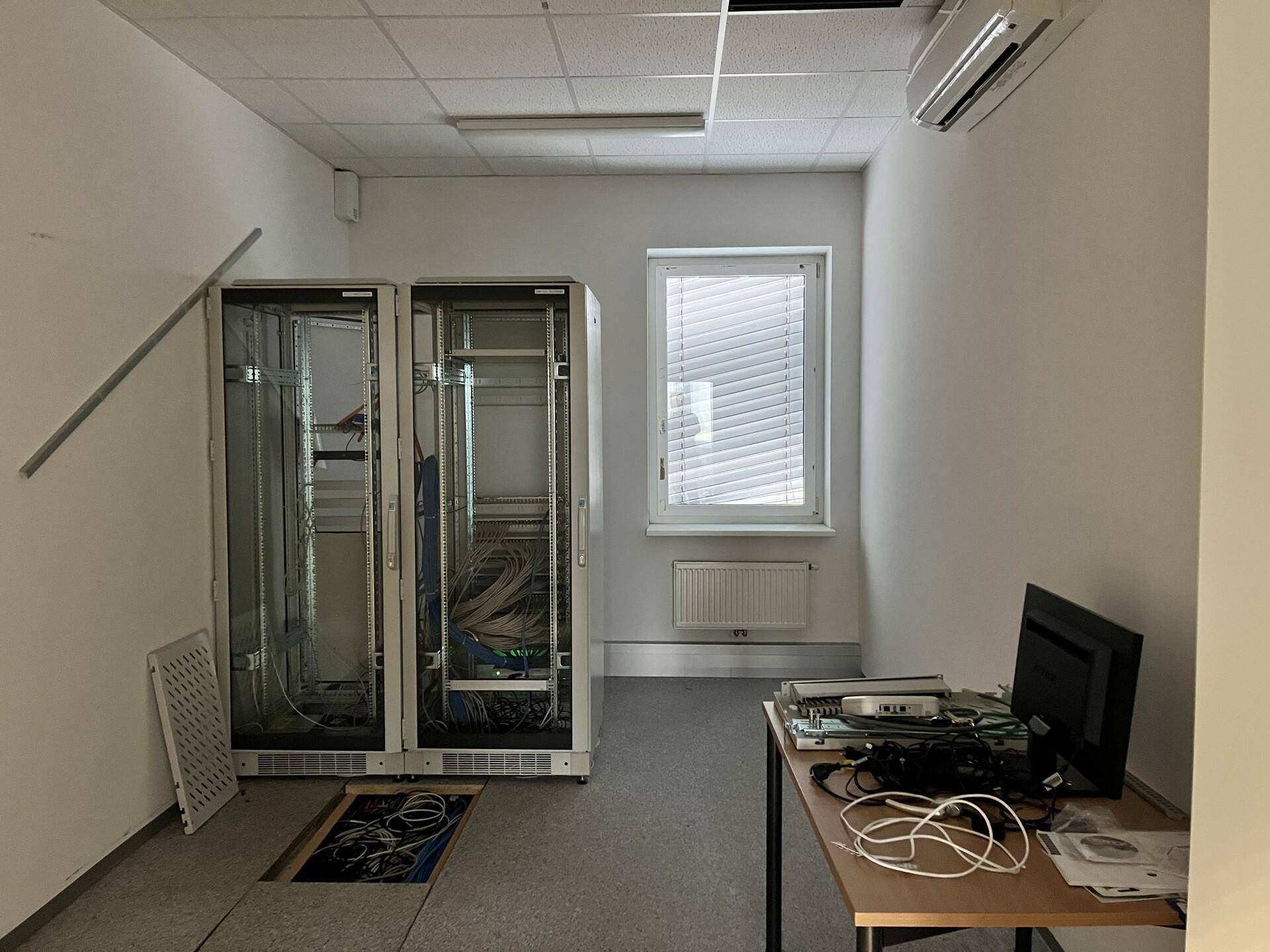 Serverraum mit zwei leeren Racks, Klimaanlage und einem Fenster mit Jalousien.