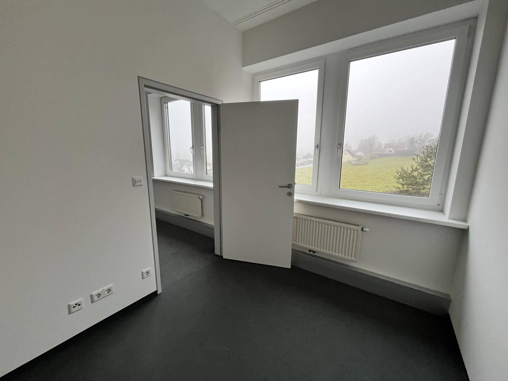 Helles Büro mit Teppichboden, Fenstern und einem Heizkörper, ideal für Einzel- oder Gruppenarbeit.
