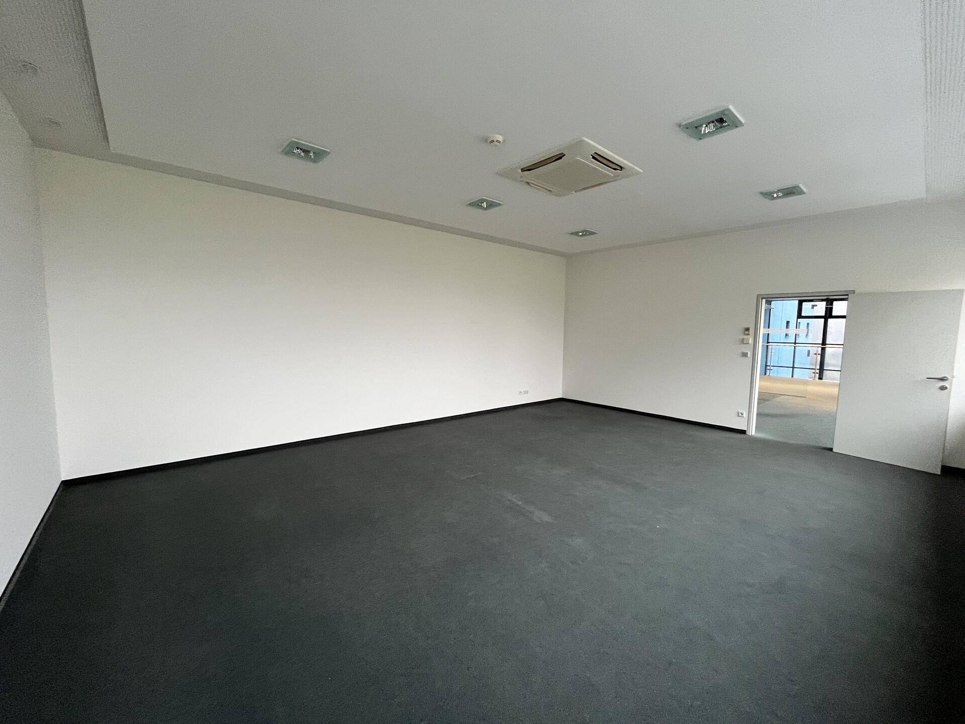 Großes, leeres Büro mit Teppichboden und einer Tür, die zu einem weiteren Bereich führt.