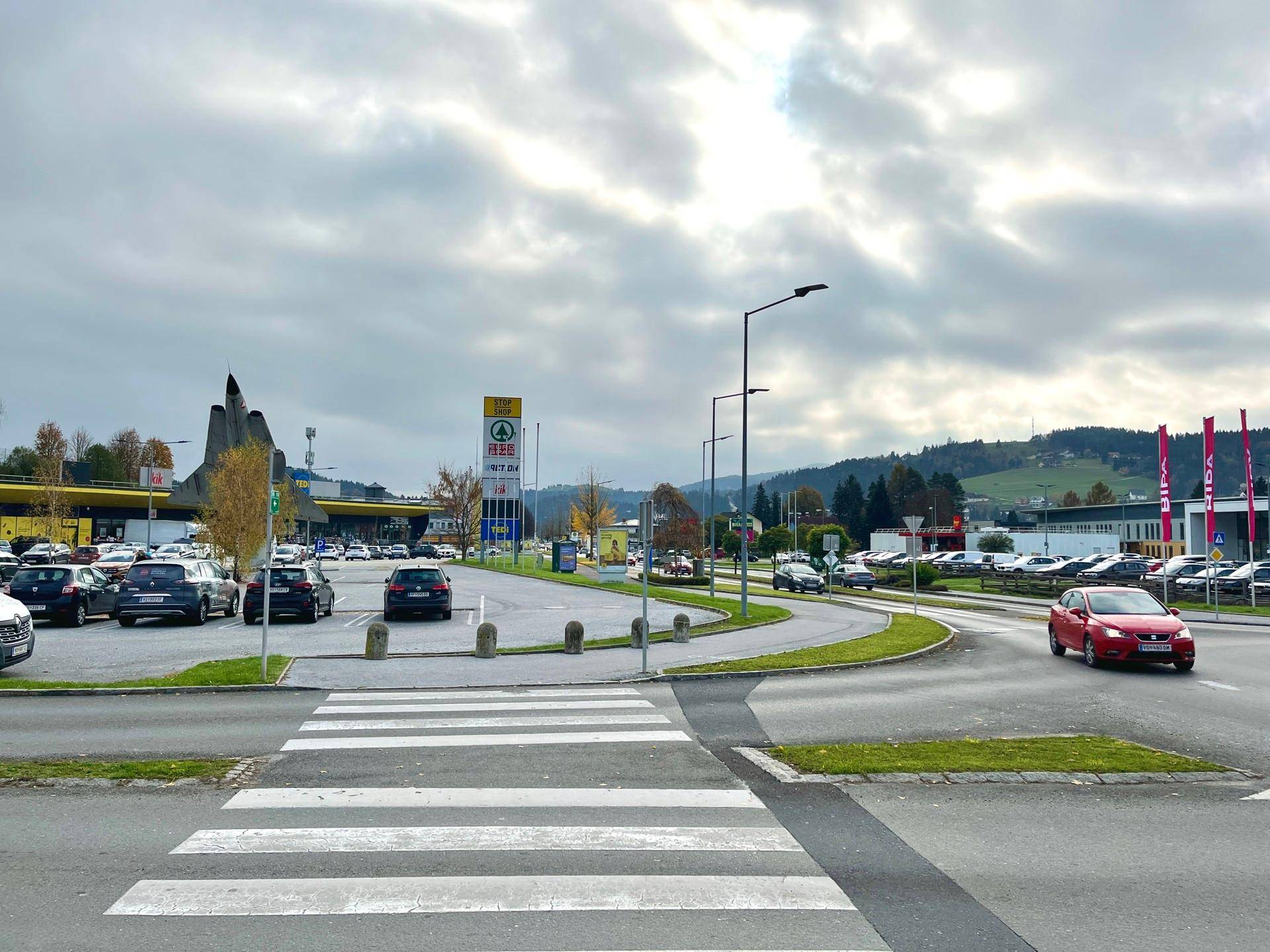Außenansicht eines Einkaufsbereichs mit Zebrastreifen, Straße und Parkplätzen unter bewölktem Himmel.