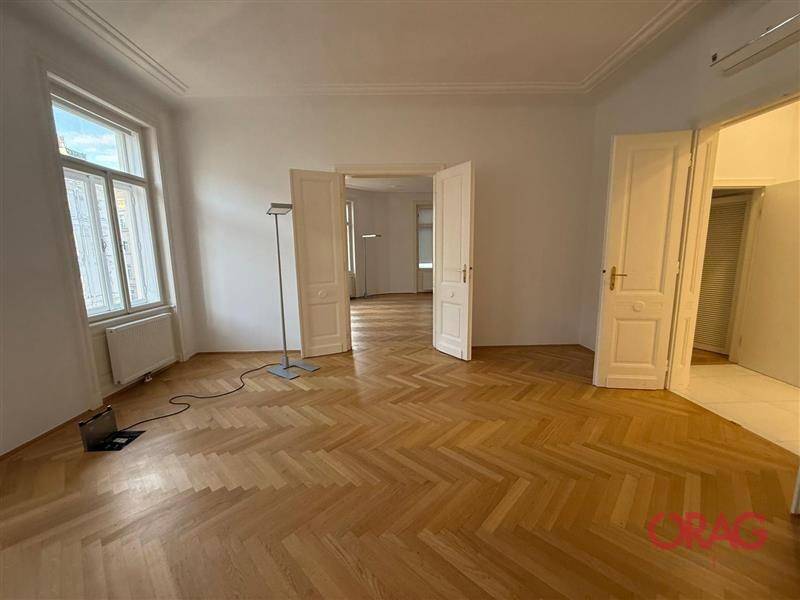 Großes, leeres Zimmer mit elegantem Fischgrätparkett, hohen Decken und viel natürlichem Licht.