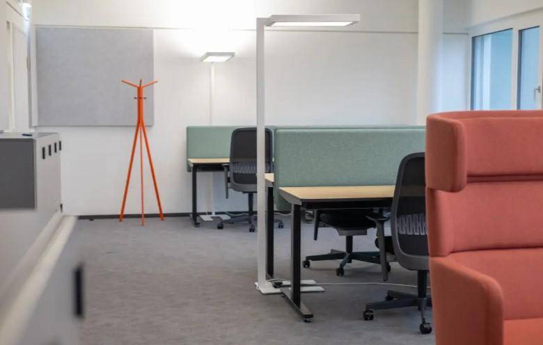 Heller Bürobereich mit mehreren Arbeitsplätzen, Trennwänden und einem auffälligen orangefarbenen Garderobenständer.