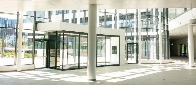 Großzügige und lichtdurchflutete Eingangshalle mit Glasfassade, modernen Säulen und viel Tageslicht.