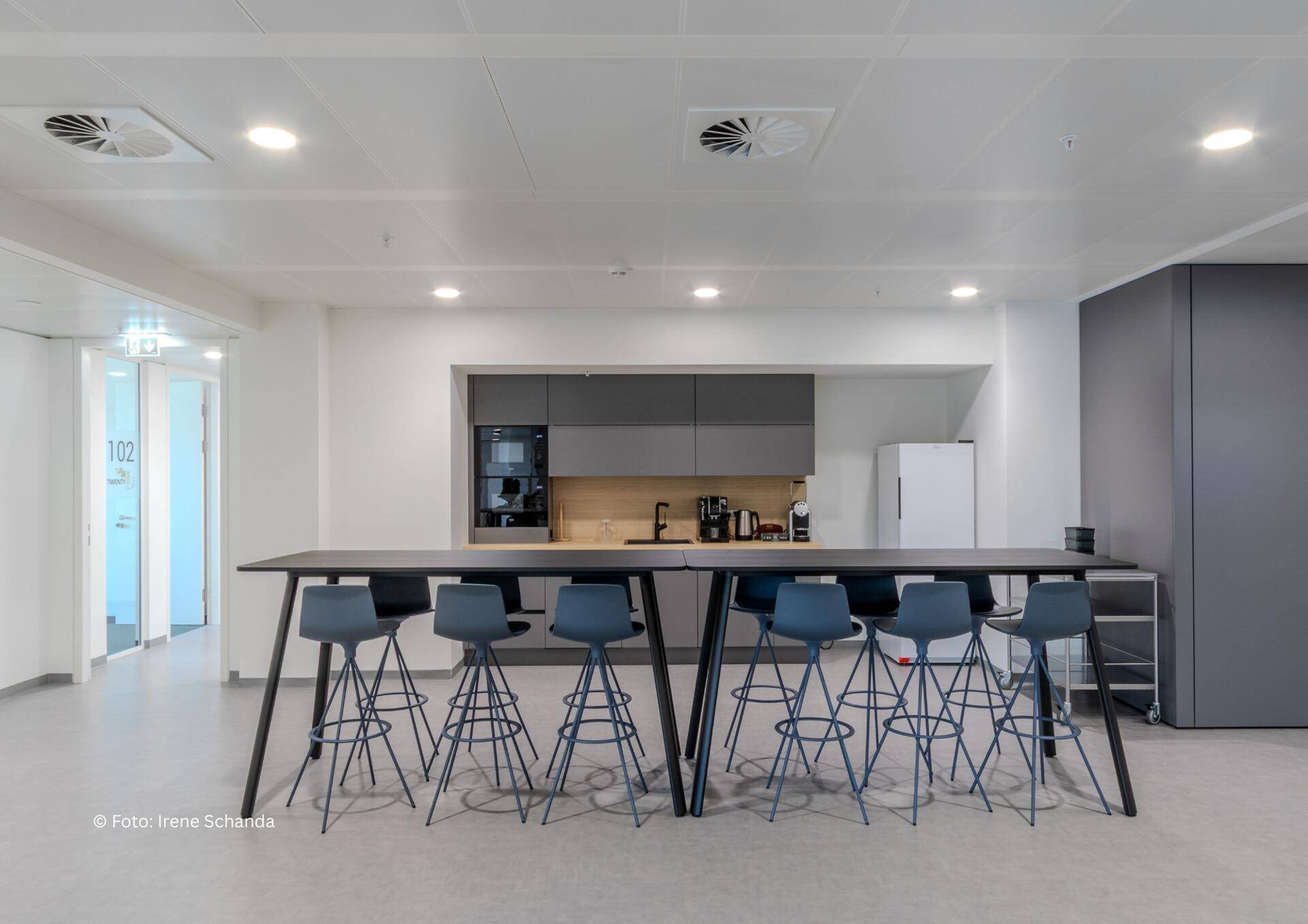 Moderne Teeküche mit langer Bar, Barhockern und integrierten Geräten in grauem Design.