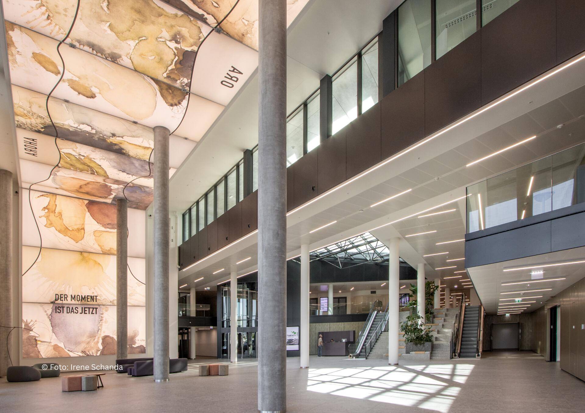 Großzügige, helle Lobby mit hohen Säulen, künstlerischer Deckenverkleidung und Treppenaufgängen in einem modernen Gebäude.