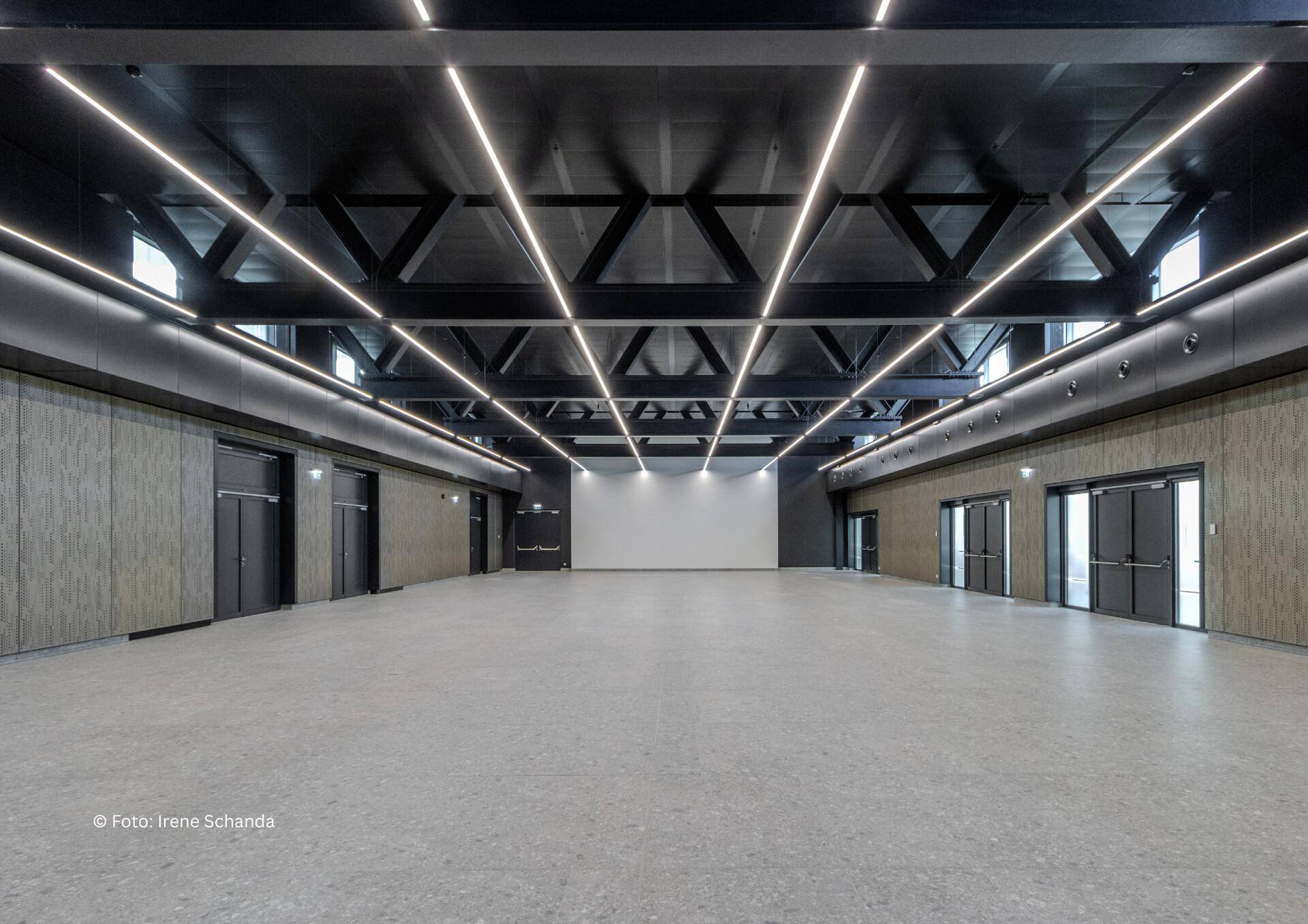 Große, leere Halle mit hoher Decke, sichtbaren Trägern und moderner Beleuchtung, ideal für Veranstaltungen.