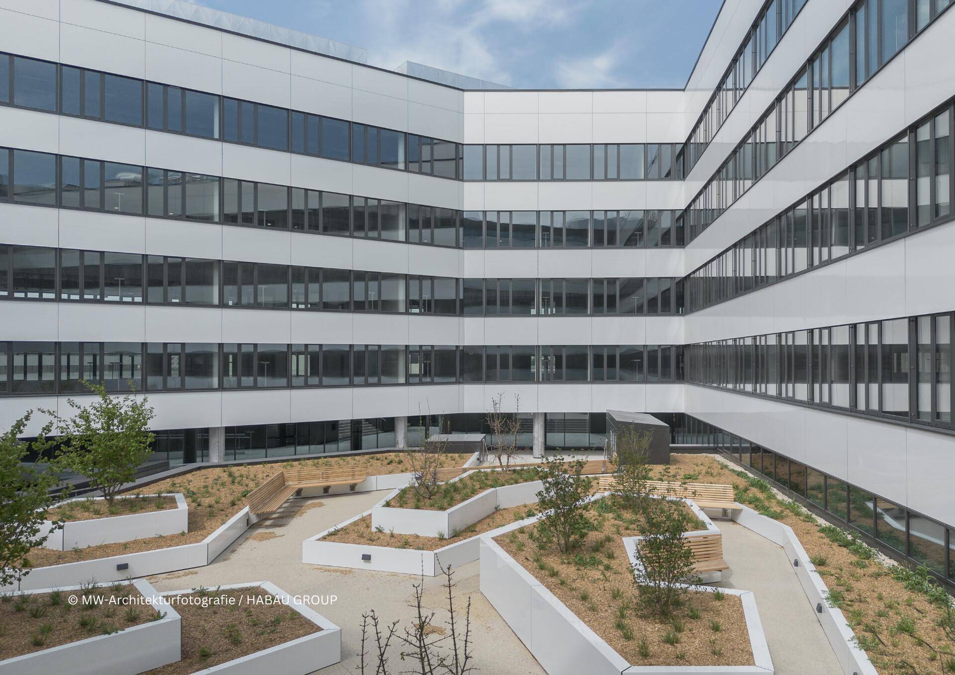 Innenhof eines modernen Bürokomplexes mit begrünten Beeten, Sitzgelegenheiten und großen Fenstern.