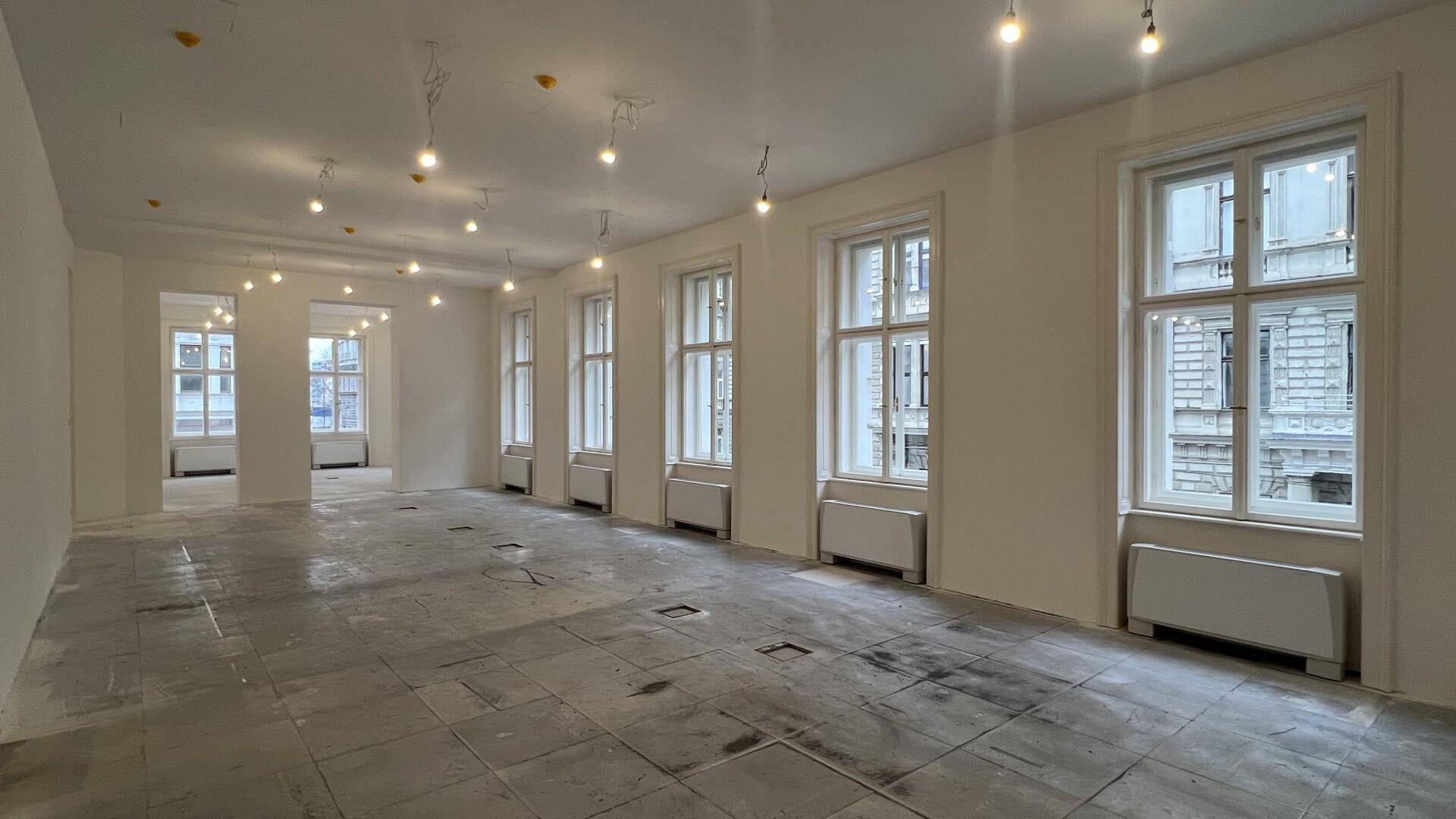 Langer, heller Büroraum mit vielen Fenstern, hohen Decken und vorbereitetem Boden, ideal für Renovierung.