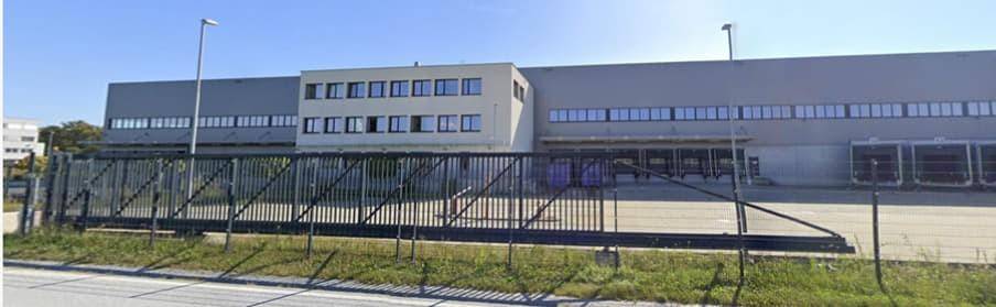 Weitläufiges Industriegebäude mit moderner Fassade, umzäuntem Gelände und Zufahrt von der Straße.