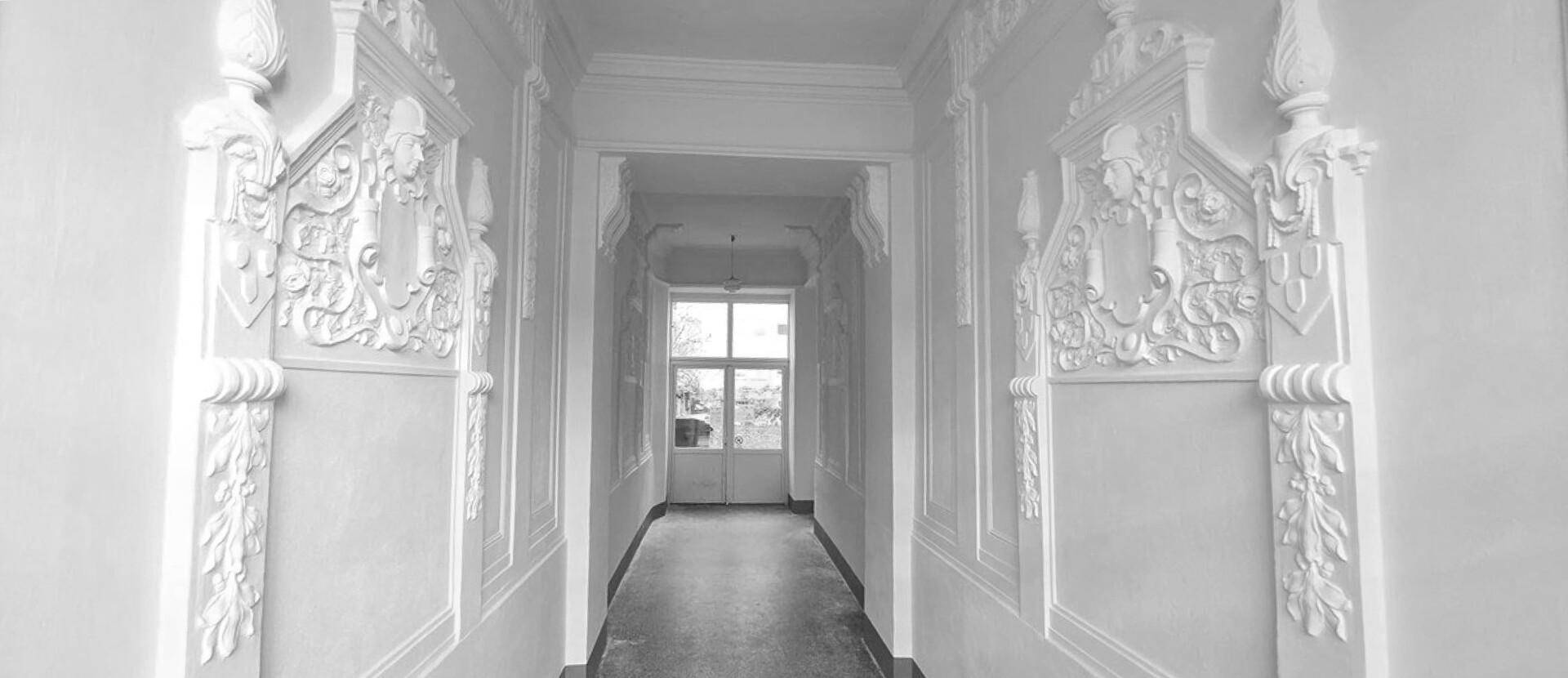Langer, eleganter Flur mit beidseitigen Stuckverzierungen und einer Tür am Ende, die Tageslicht hereinlässt.