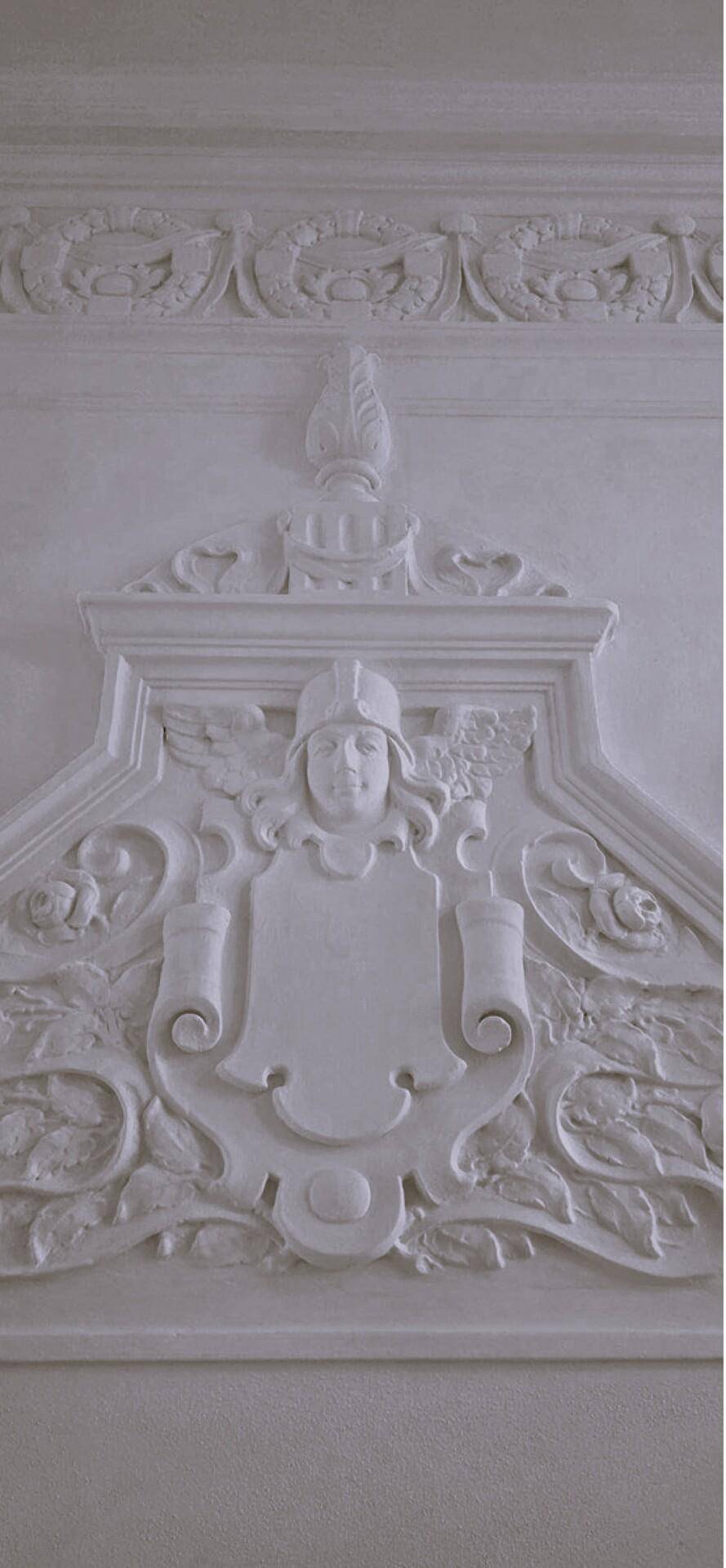 Detailaufnahme einer aufwendigen Stuckverzierung an einer Innenwand mit heraldischen Elementen.