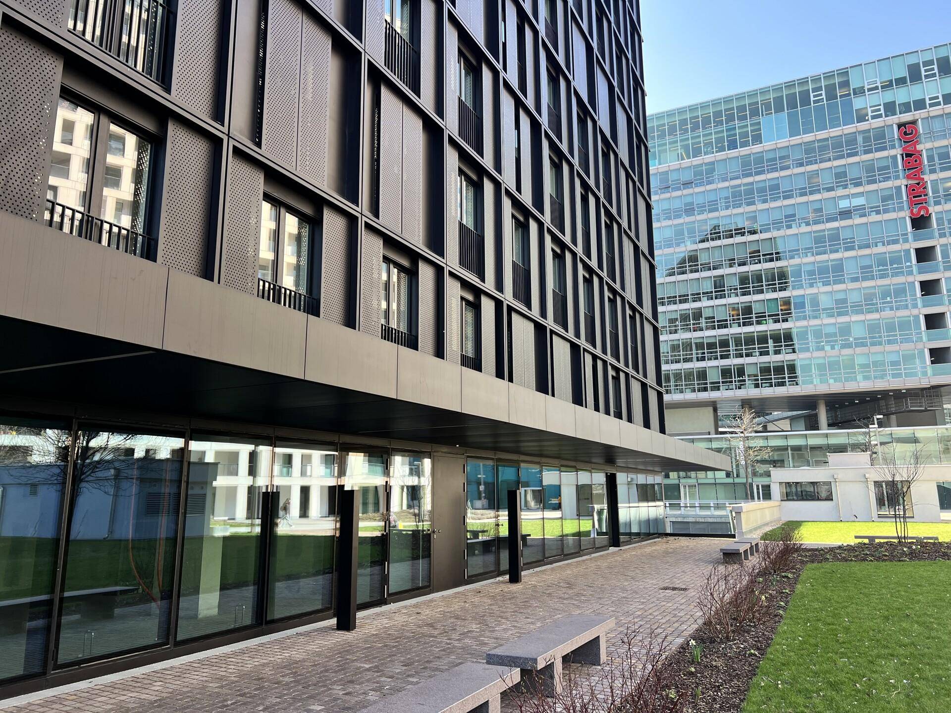 Moderne Gebäudefassade mit großen Glasflächen im Erdgeschoss und angrenzender Grünfläche.