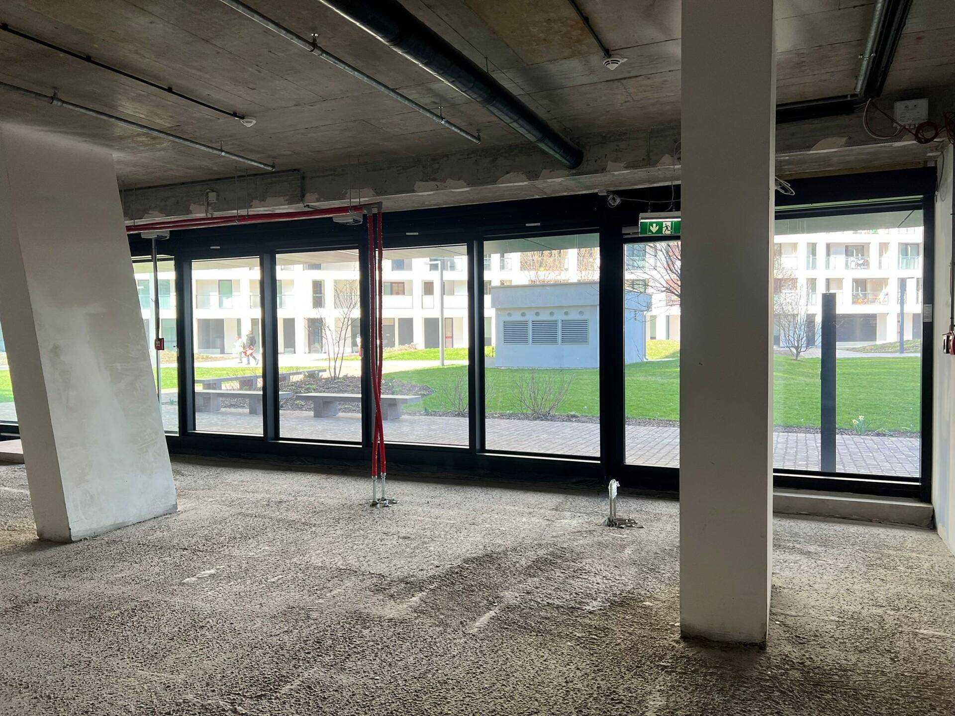 Unfertiger Gewerberaum mit großen Fenstern und Blick auf eine Grünfläche, gekennzeichnet durch eine rohe Betonoberfläche und sichtbare Installationen.