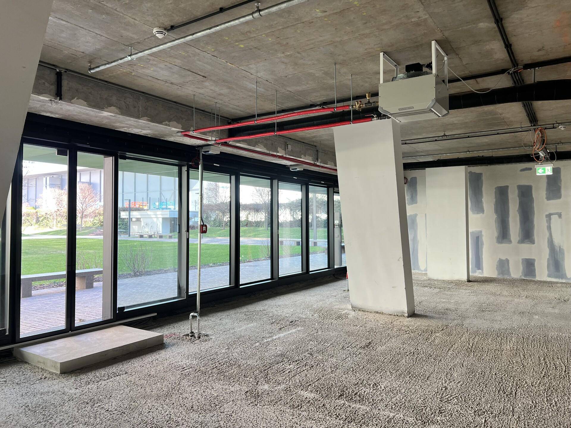 Unfertiger Gewerberaum mit großen Fenstern, Blick ins Grüne und sichtbaren Deckeninstallationen und Lüftungselementen.