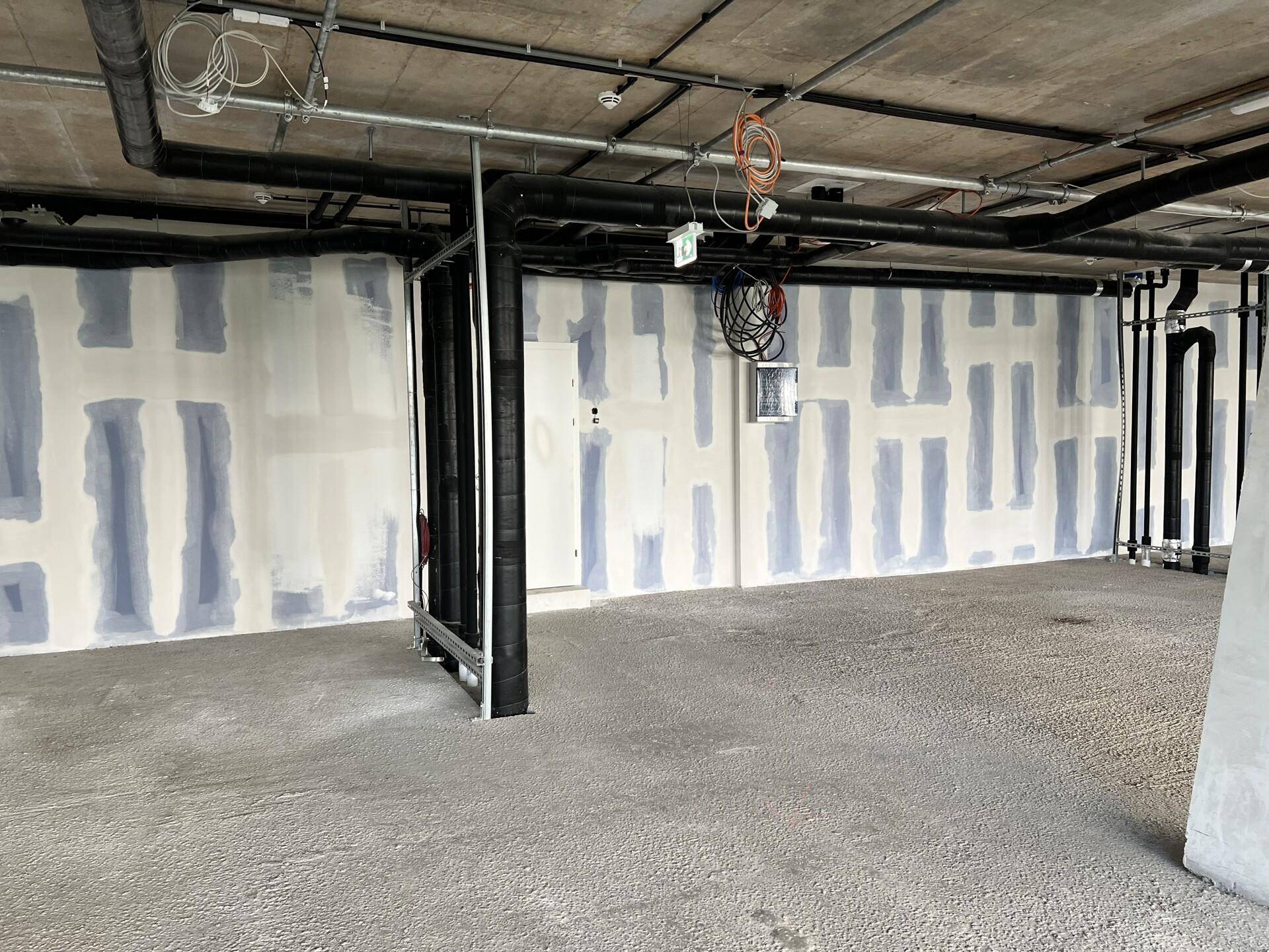 Unfertiger Gewerberaum mit teilweise verputzten Wänden, sichtbaren Rohren und einem rohen Betonboden.