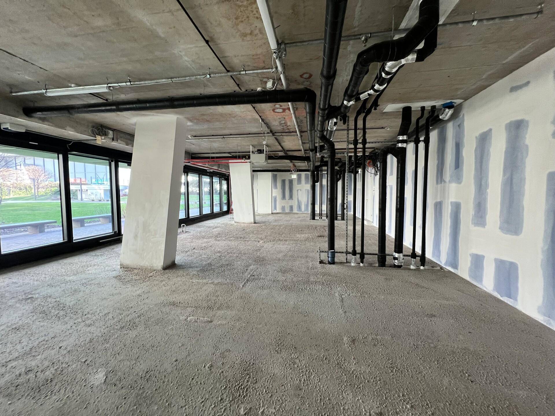 Großer, unfertiger Gewerberaum mit sichtbaren Rohren an der Decke und Wänden sowie einem rohen Betonboden.
