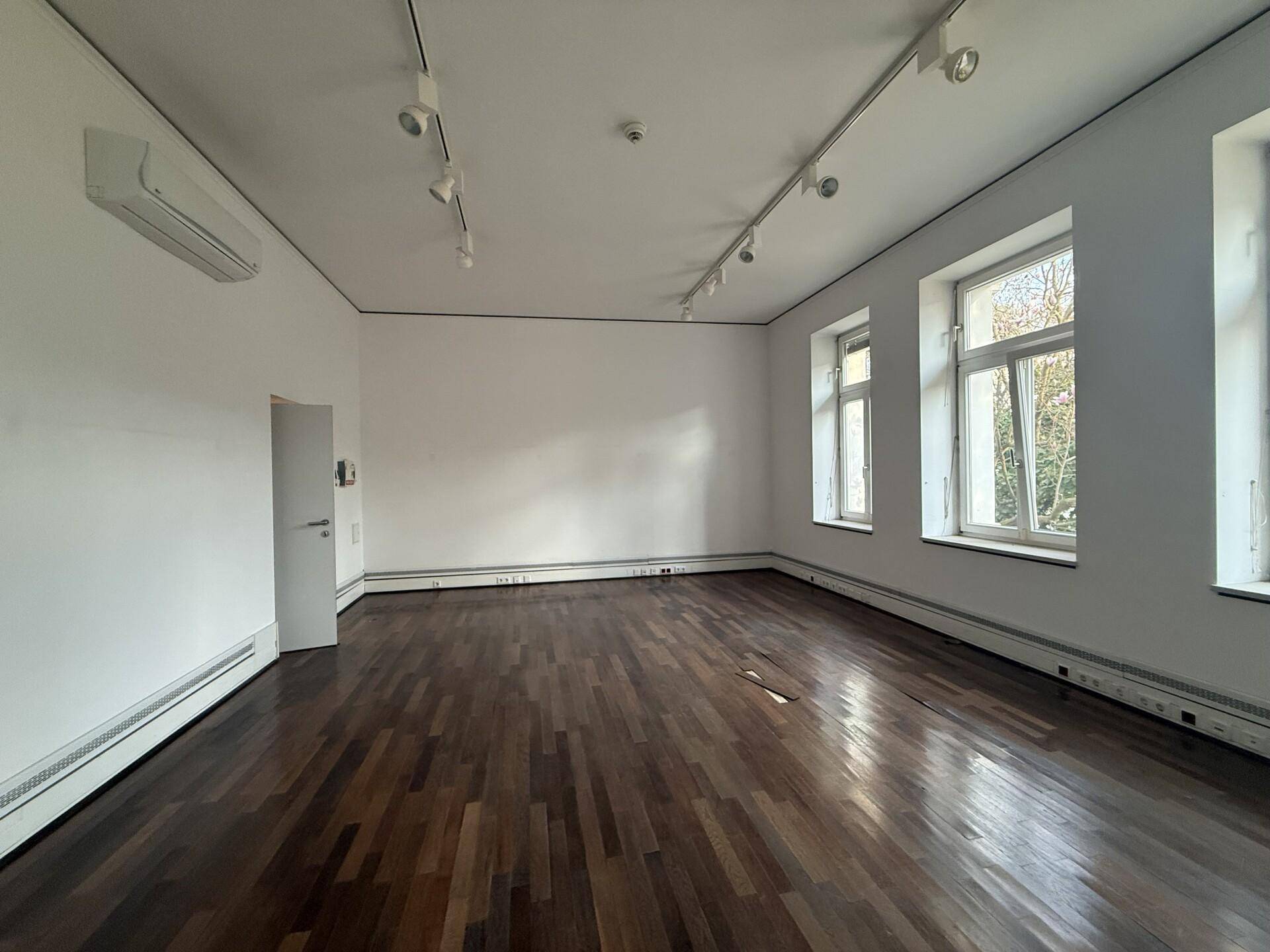 Heller, leerer Raum mit dunklem Parkettboden, großen Fenstern und einer Klimaanlage an der Wand.