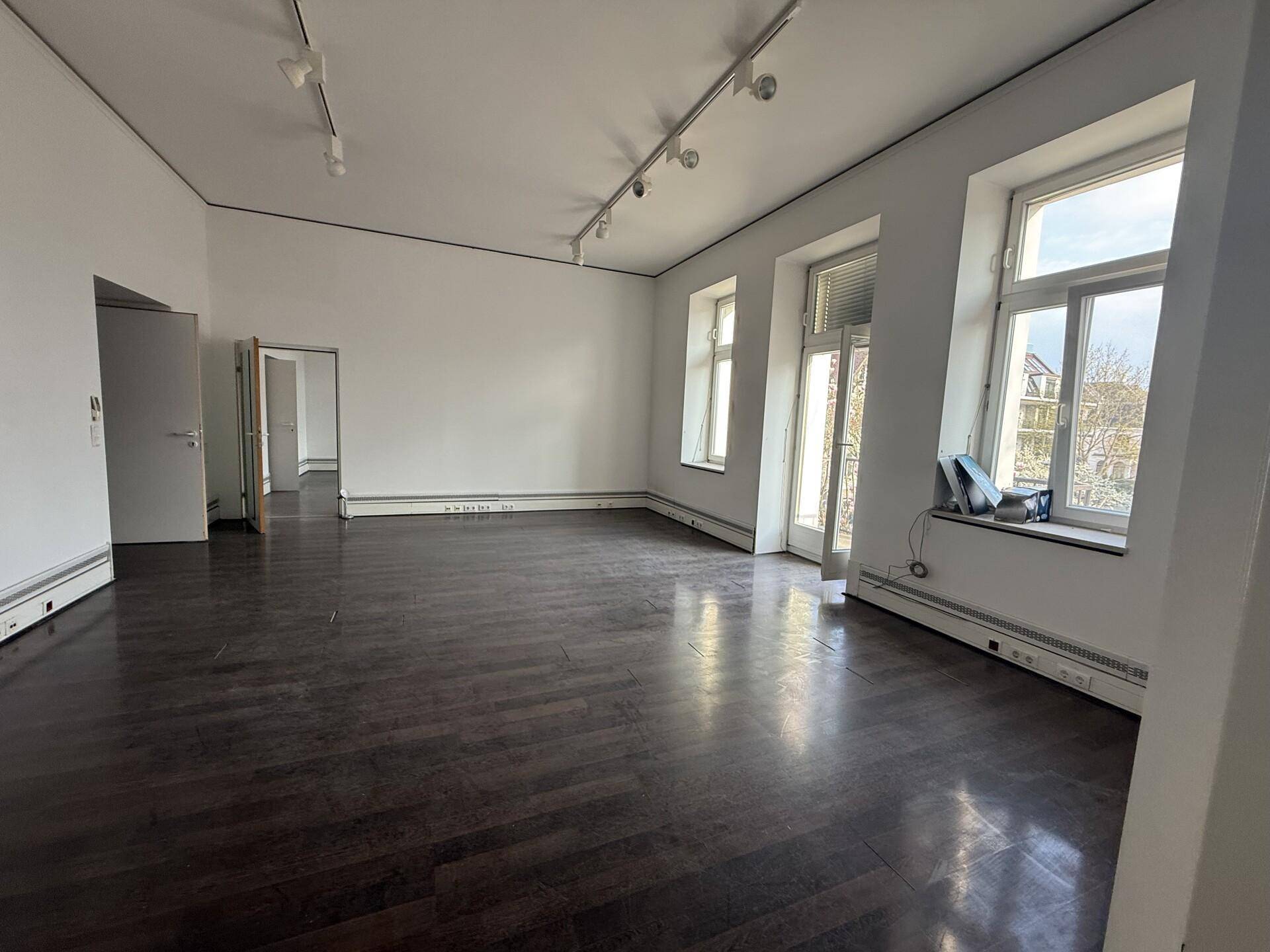 Großer, heller Raum mit dunklem Holzboden, hohen Decken und Zugang zu einem kleinen Balkon.