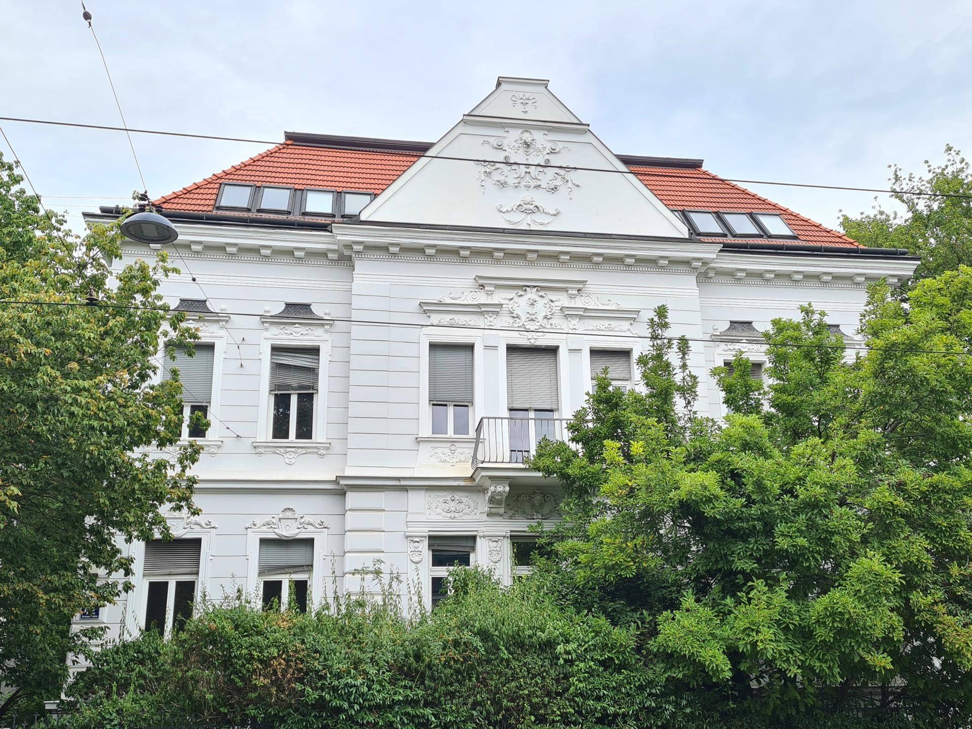 Klassisches Gebäude mit rotem Ziegeldach, weißer Fassade und üppigen grünen Bäumen im Vordergrund.