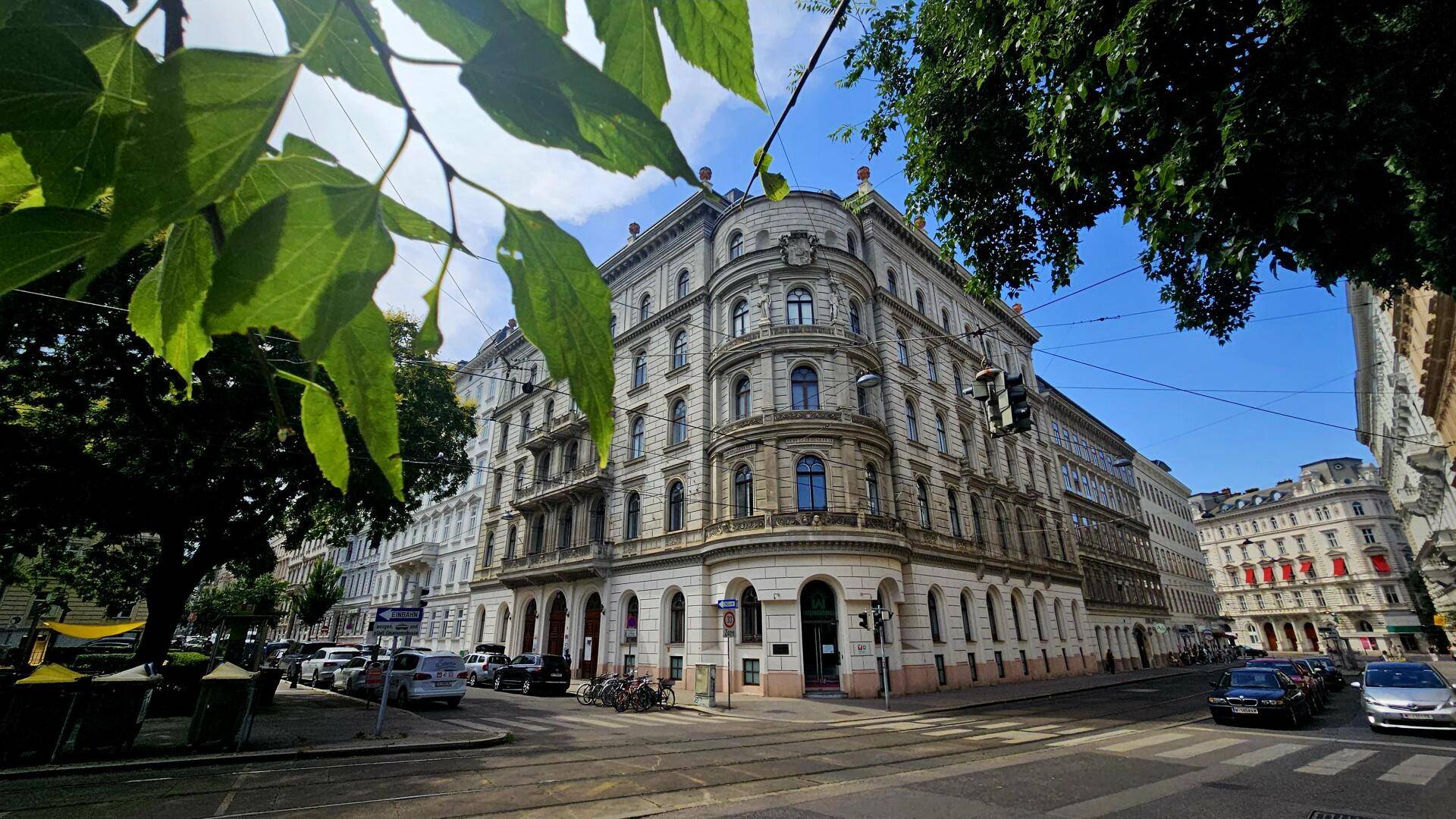 Historisches Gebäude an einer belebten Straßenecke mit vielen Fenstern und grünen Bäumen.