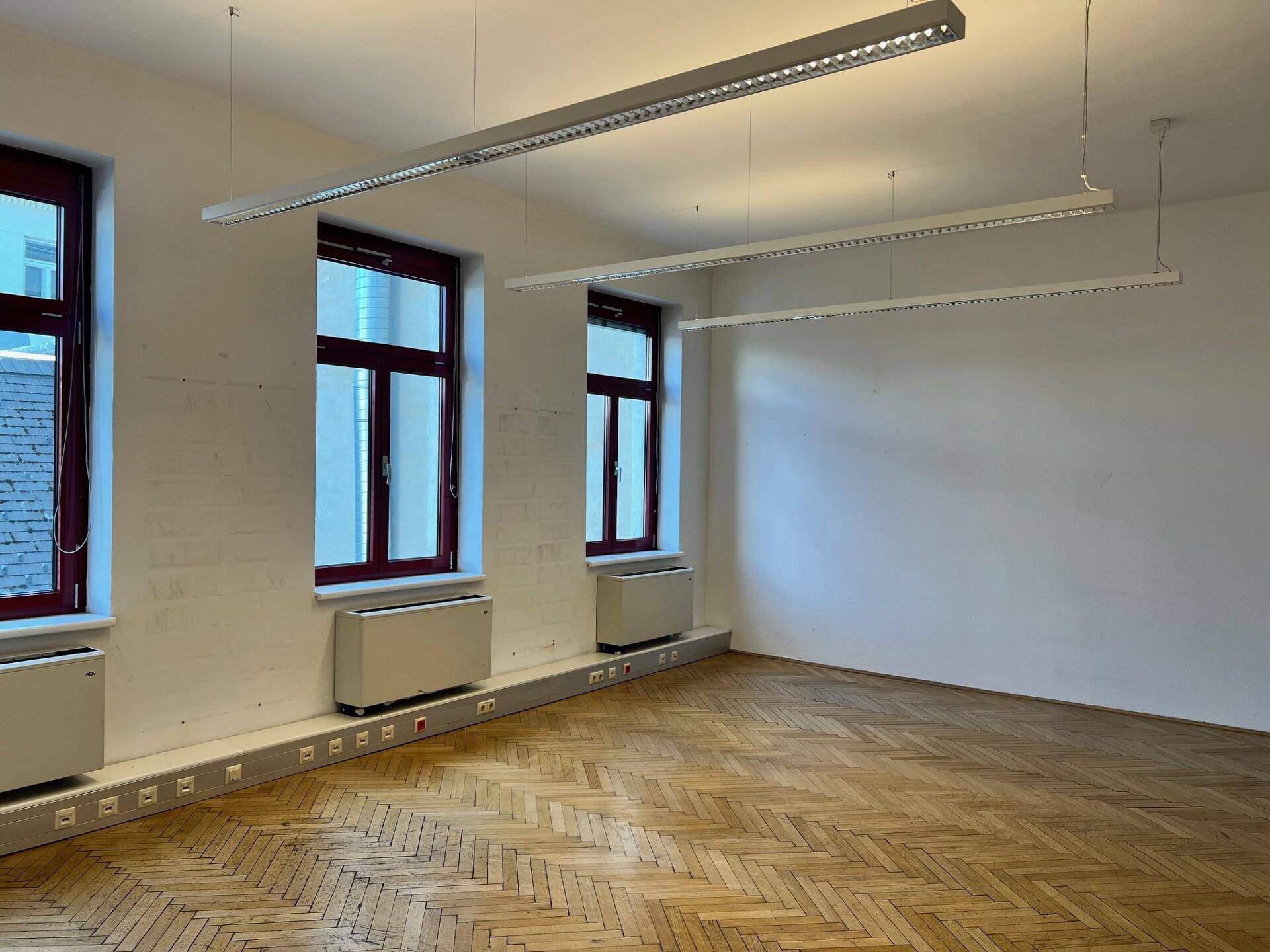 Geräumiges Büro mit Fischgrätparkett, großen Fenstern und modernen Deckenleuchten.
