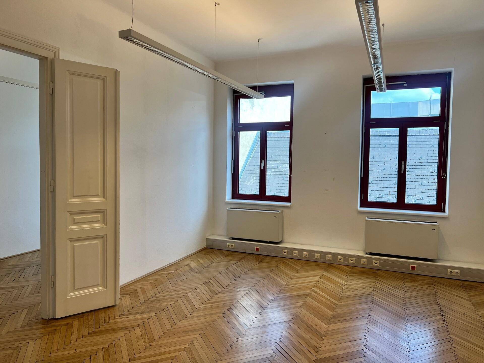 Leeres Büro mit Fischgrätparkett, hohen Decken und großen Fenstern, die viel Licht hereinlassen.