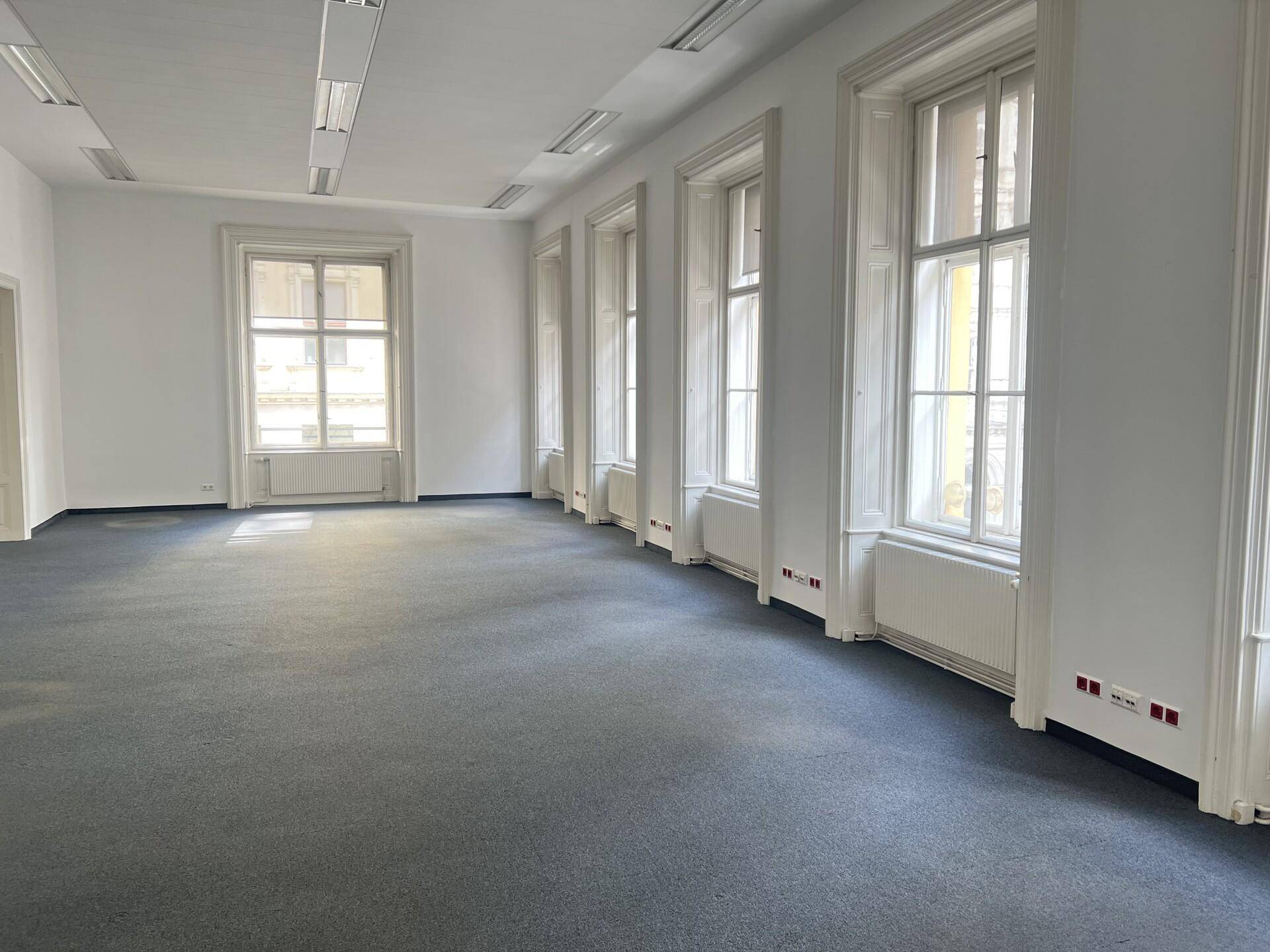 Großer, heller Büroraum mit grauem Teppichboden, hohen Fenstern und viel Tageslicht.