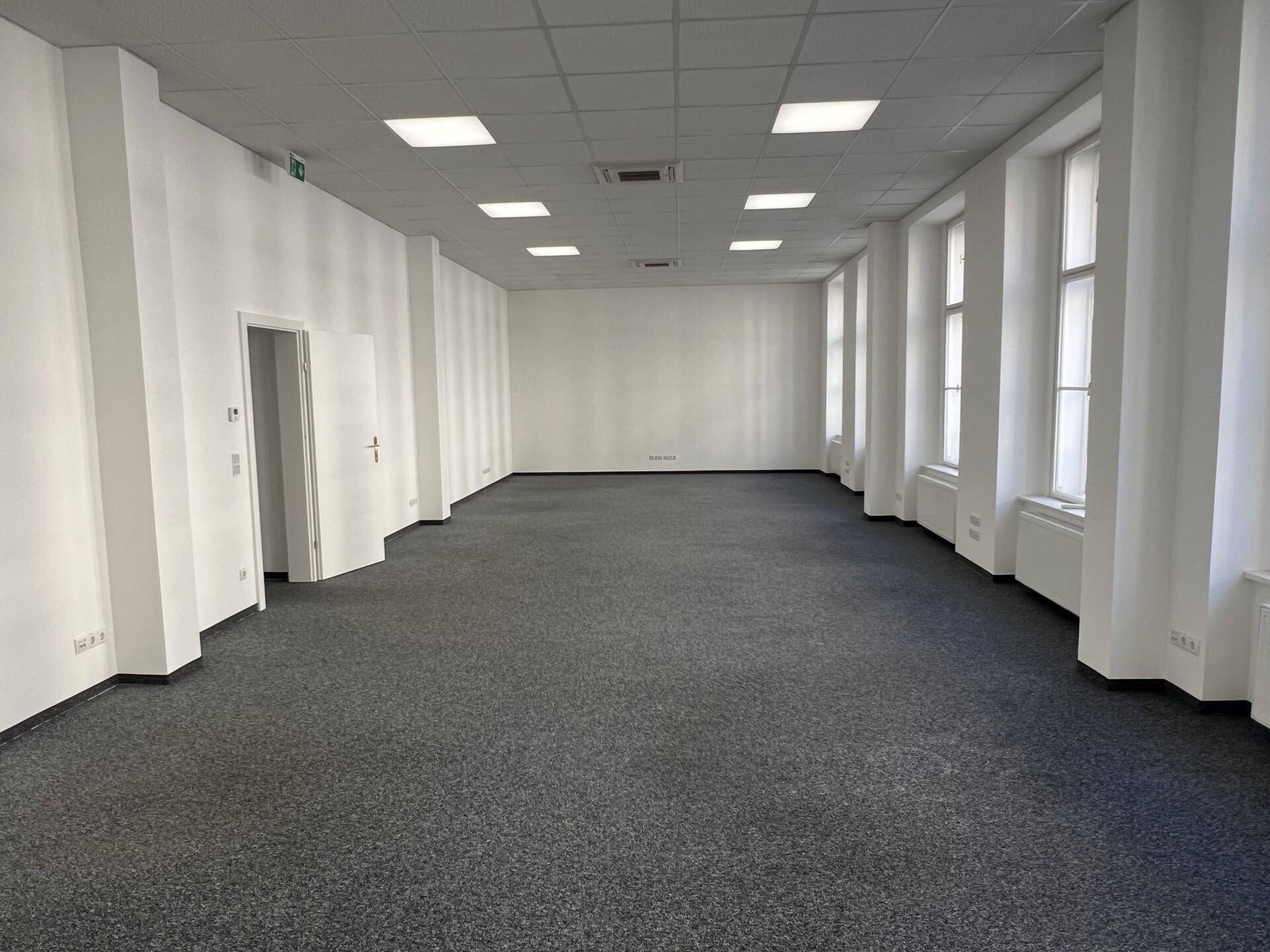 Geräumiger, leerer Büroraum mit grauem Teppichboden, hohen Fenstern und moderner Deckenbeleuchtung.