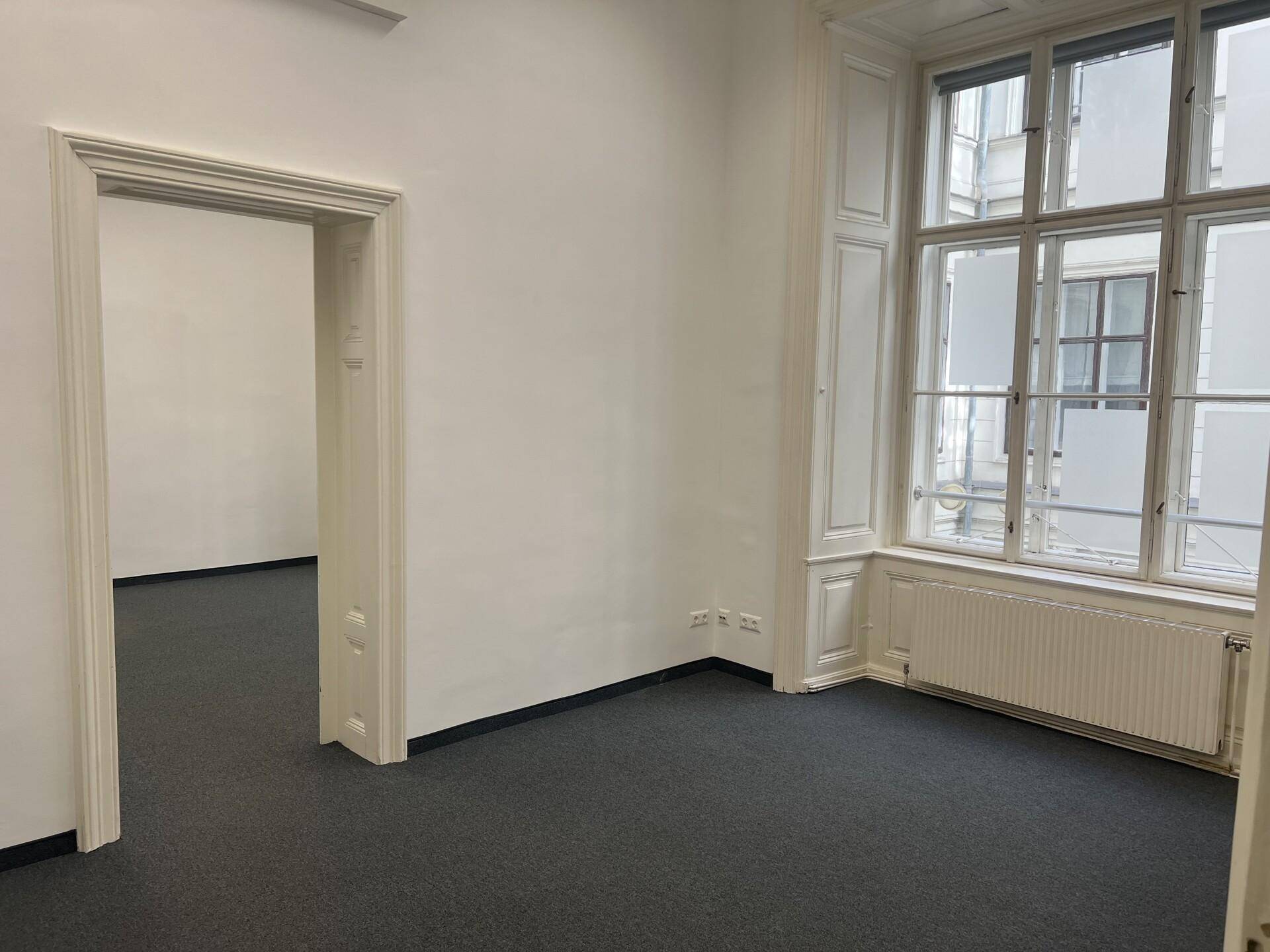 Heller Büroraum mit Teppichboden, hohen Fenstern und klassischer Wandverkleidung.