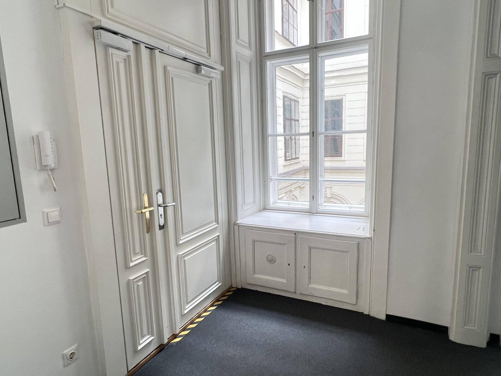 Heller Eingangsbereich mit klassischer Holztür, Fenster und integriertem Schrank unter der Fensterbank, mit Teppichboden.