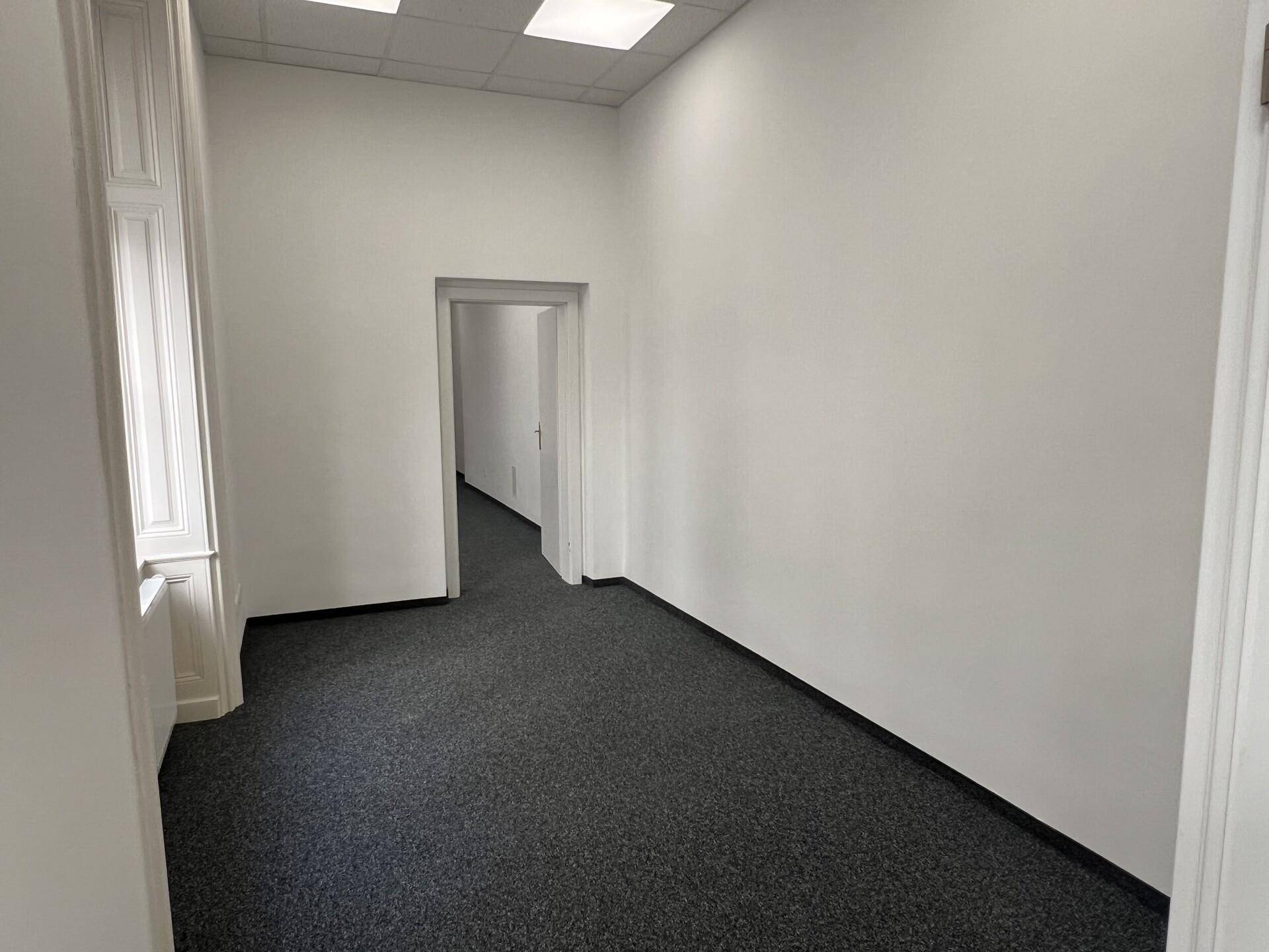 Leerer Büroraum mit grauem Teppichboden, weißen Wänden und einem Durchgang zu einem weiteren Bereich.