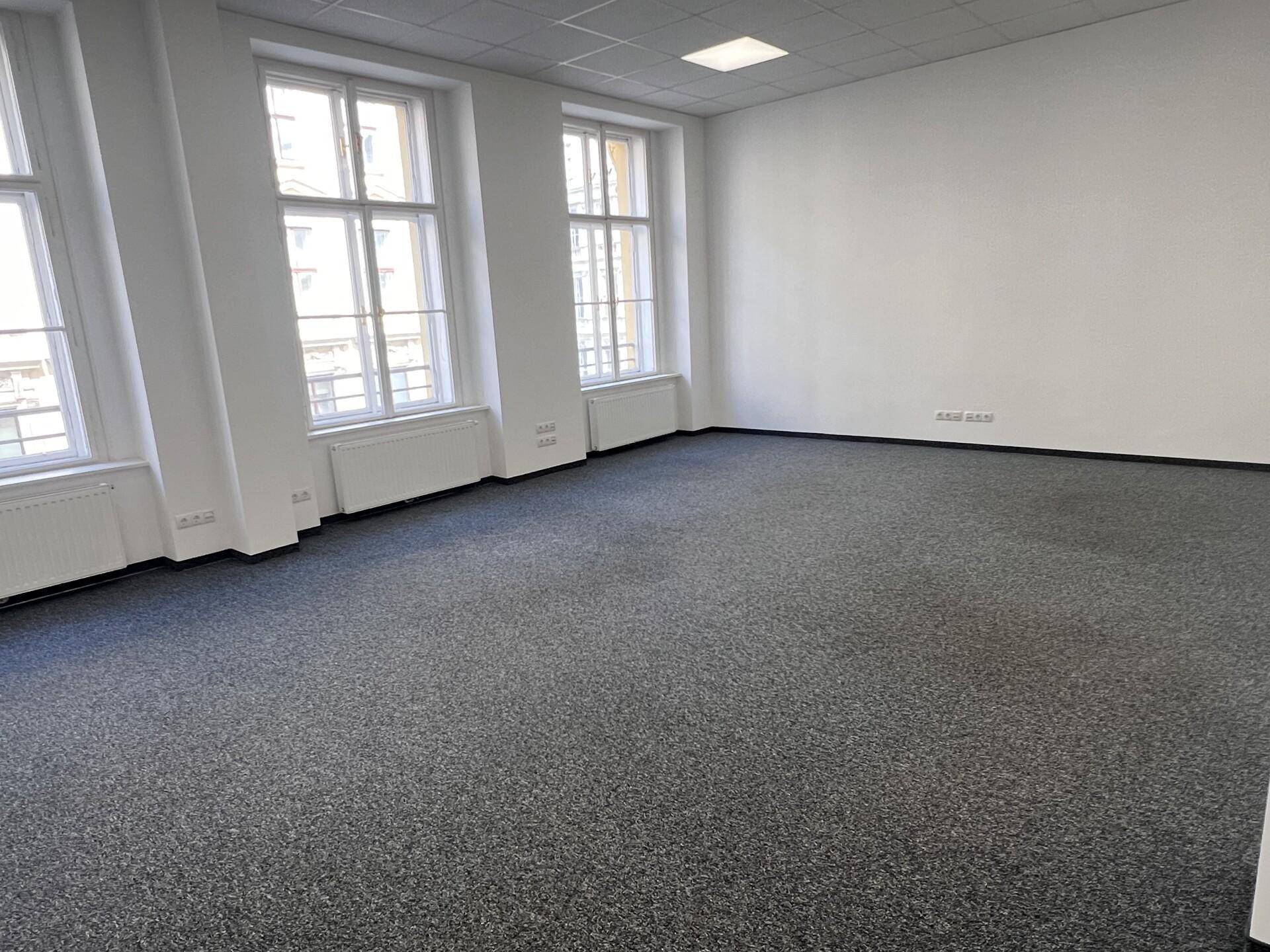 Heller Büroraum mit grauem Teppichboden, großen Fenstern und weiß gestrichenen Wänden.