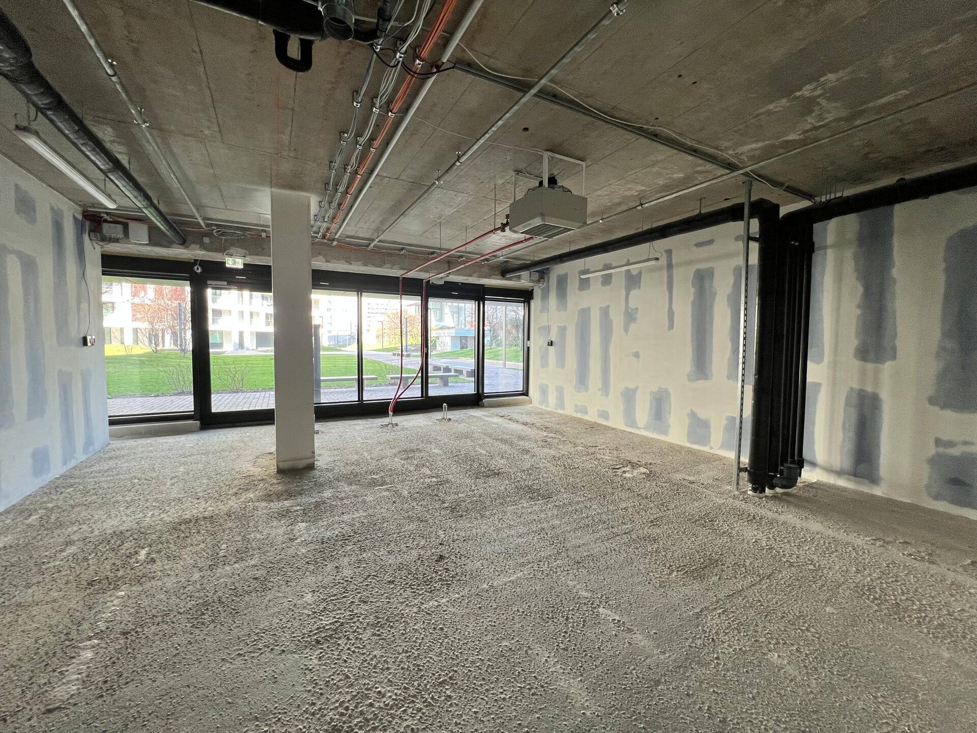 Unfertiger Innenraum mit Betonboden, sichtbaren Leitungen an der Decke und großen Fenstern mit Blick ins Grüne.