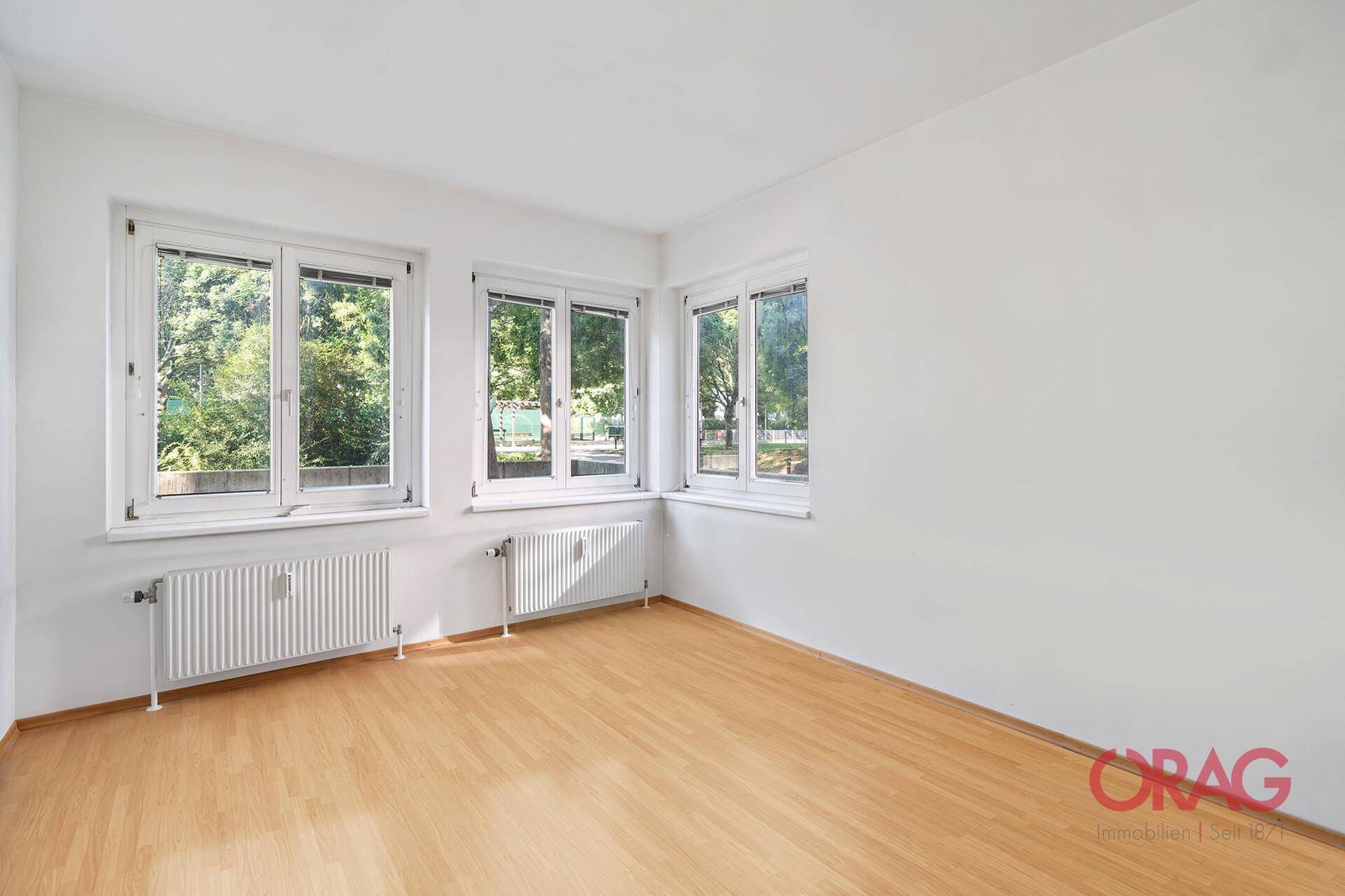 Leeres Zimmer mit hellem Laminatboden, weißen Wänden und Fenstern mit Blick auf grüne Bäume.