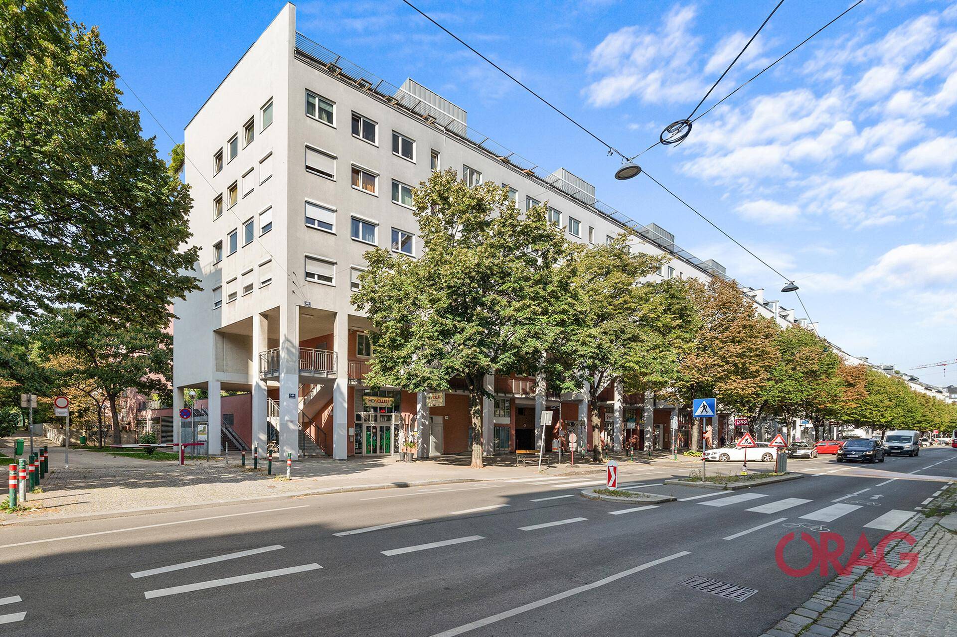 Moderne Wohnanlage mit mehreren Etagen und Bäumen entlang der Straße unter blauem Himmel.