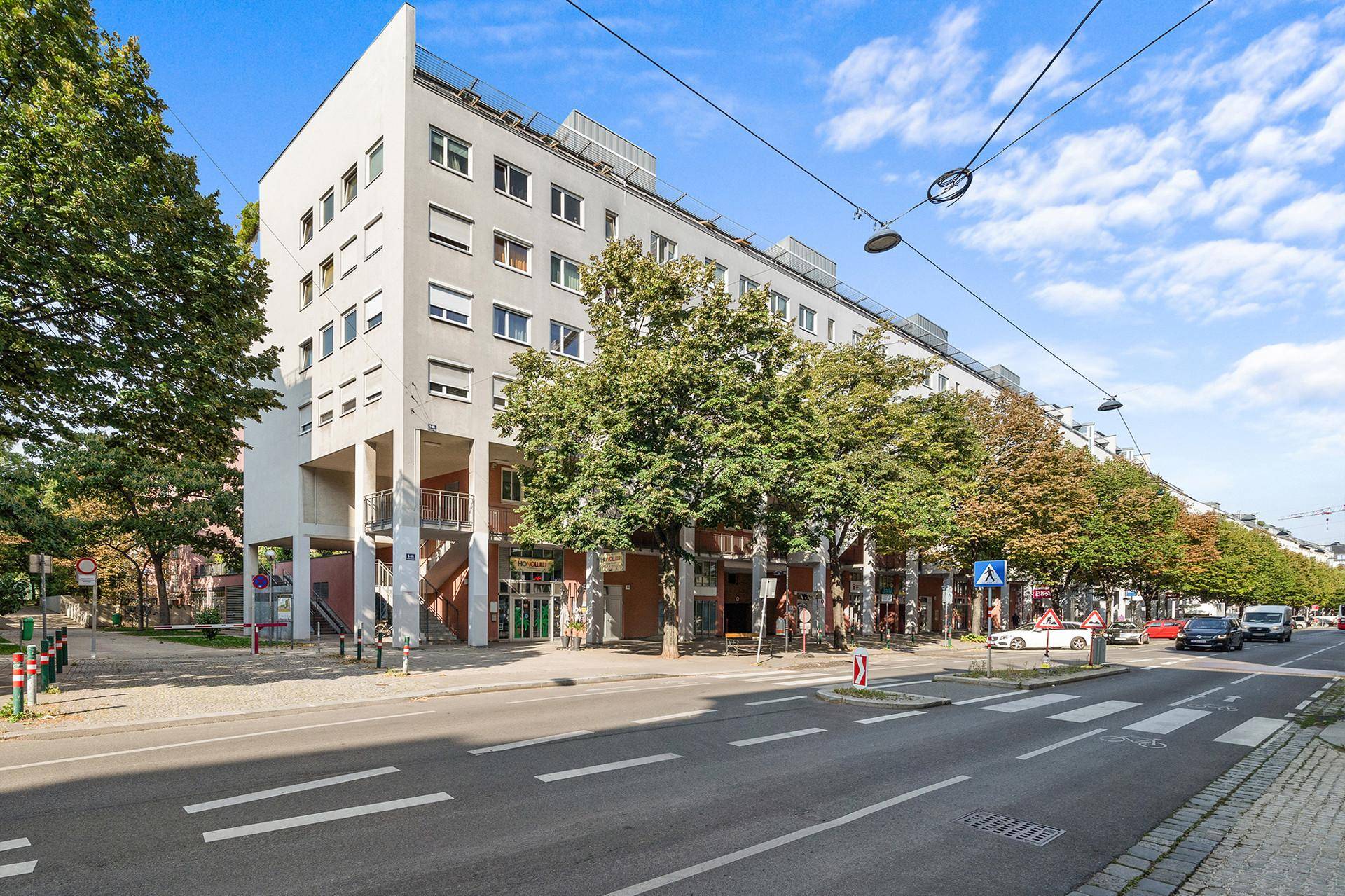 Modernes Wohngebäude mit mehreren Etagen, großen Fenstern und Bäumen entlang einer belebten Stadtstraße unter blauem Himmel.