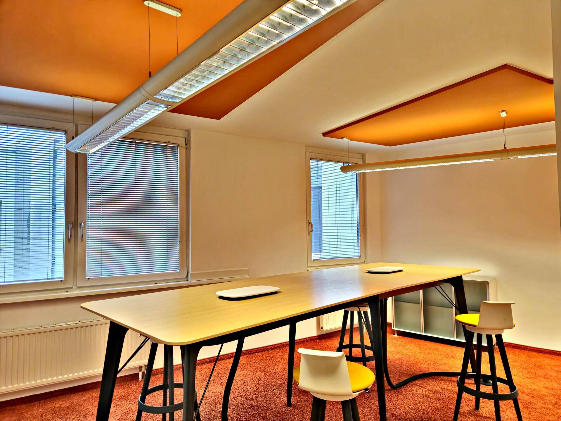 Kreativer Büroraum mit langer Holztafel, orangefarbener Decke und passendem Teppichboden, beleuchtet von modernen Hängelampen.